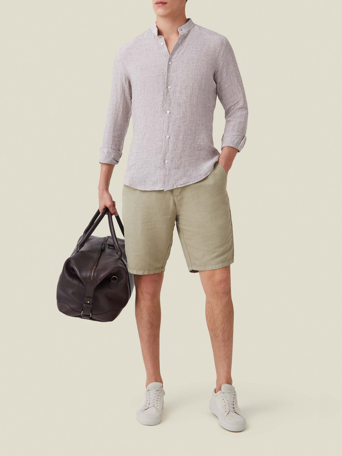Nocciola brown striped Versilia linen shirt with a band collar, styled with green shorts and a brown leather bag.