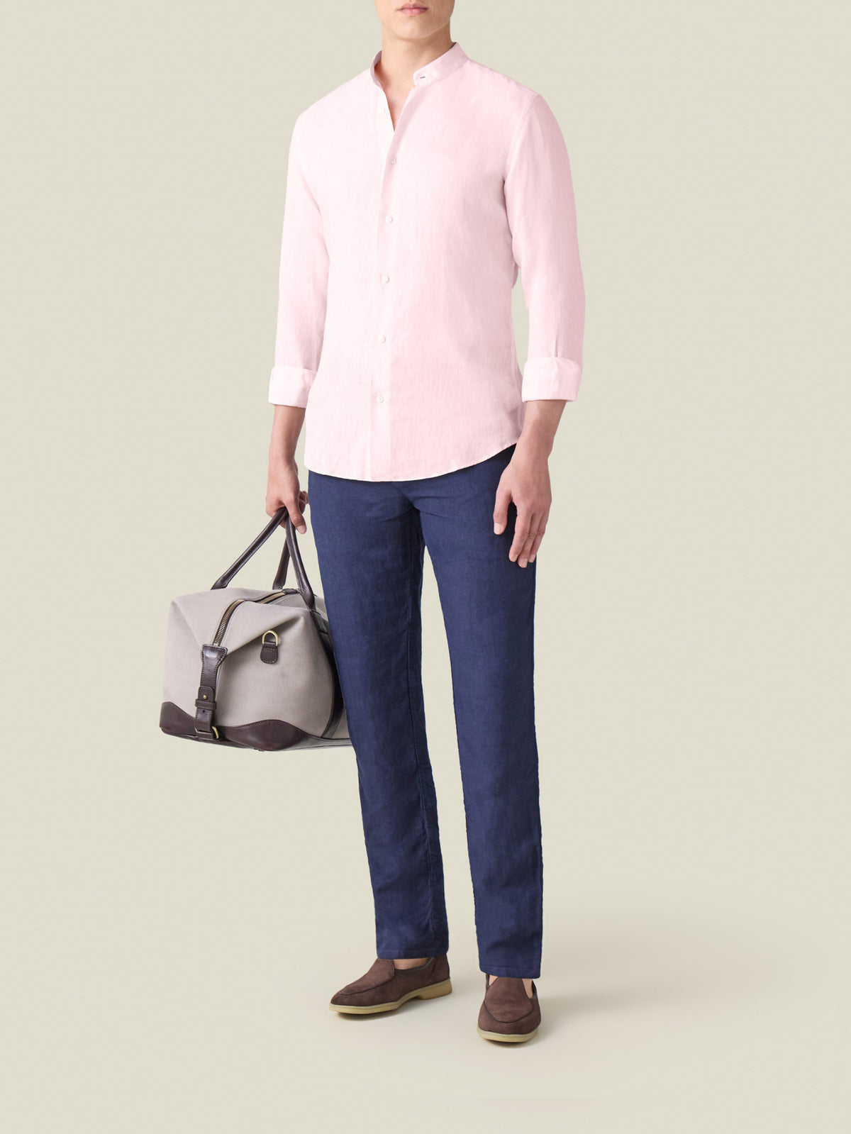 Light pink Versilia Linen Shirt with a band collar, styled with navy blue trousers and a grey weekend bag.