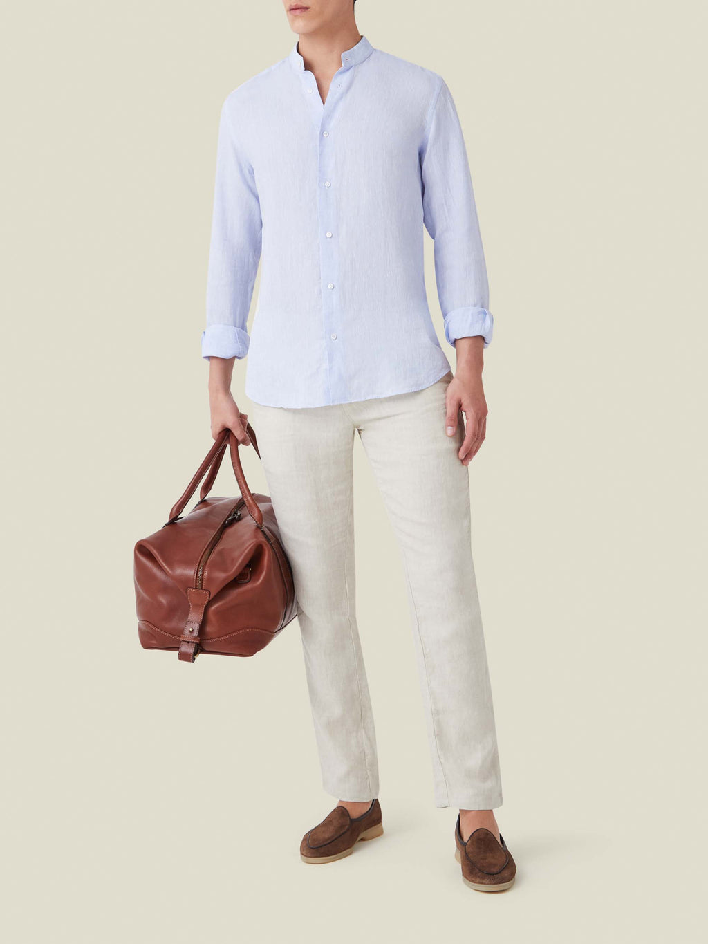Sky blue Versilia linen shirt with a band collar, worn with light trousers and a brown leather weekender bag.