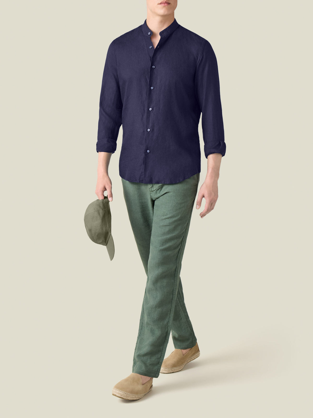 Man wearing a midnight blue linen shirt with a band collar, green trousers, and holding a matching cap.