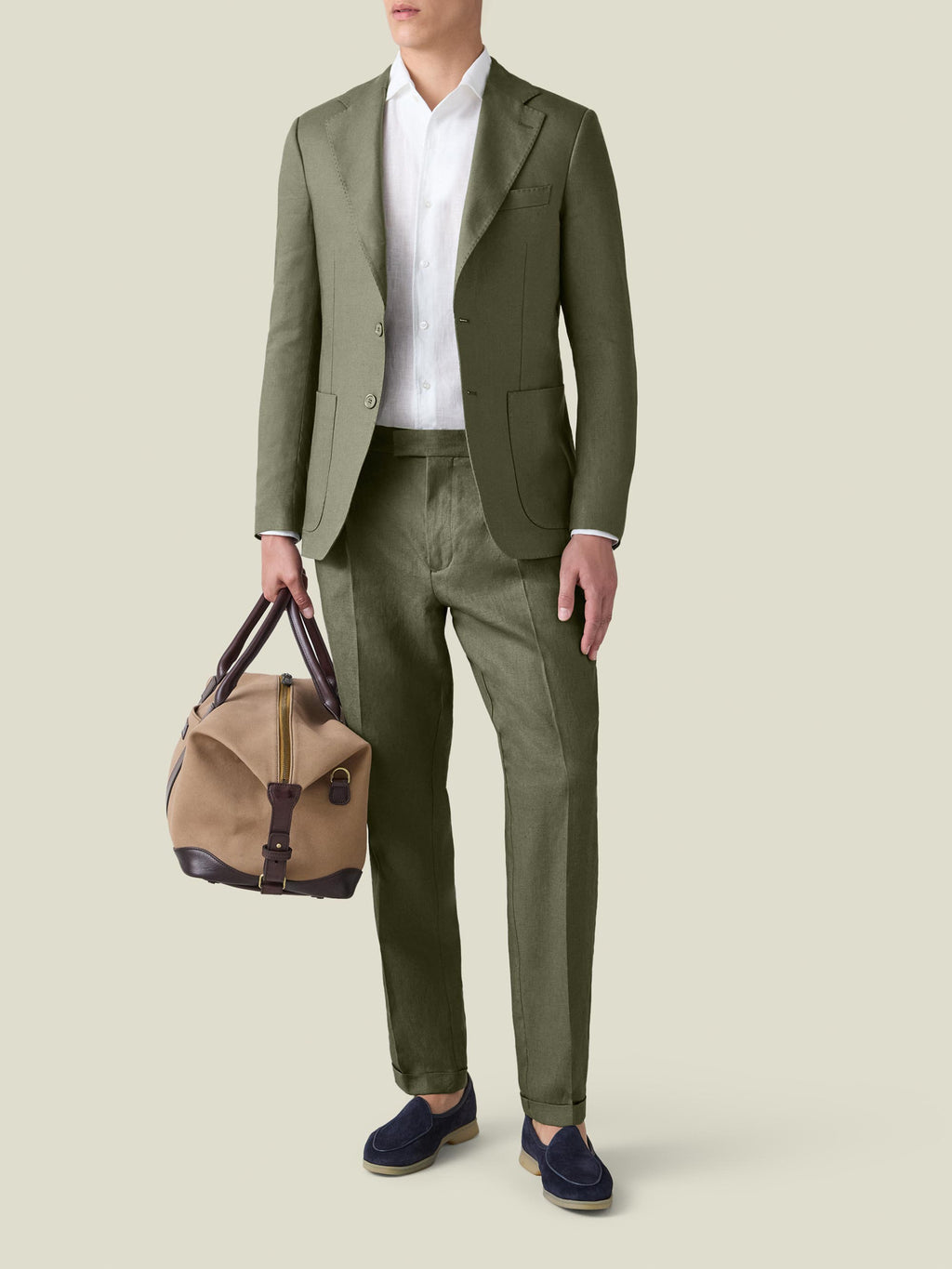 A model wears the Luca Faloni Linen Suit in Olive Green, styled with a white shirt, navy loafers, and a travel bag.