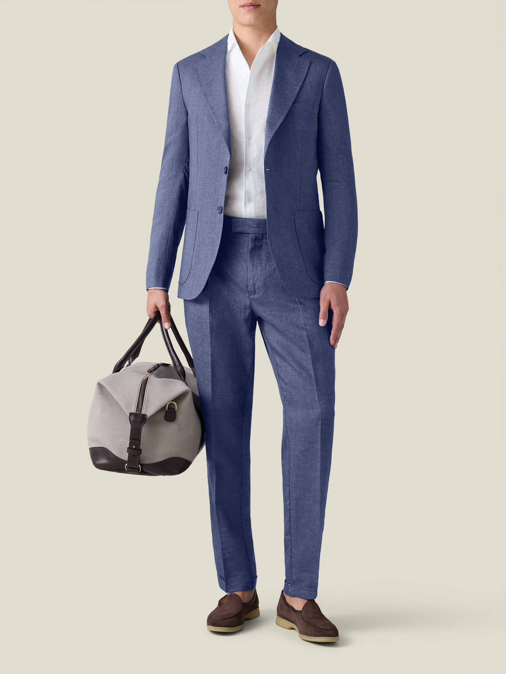 A man wears the Azure Linen Blazer and matching trousers with a white shirt, holding a grey and brown travel bag.
