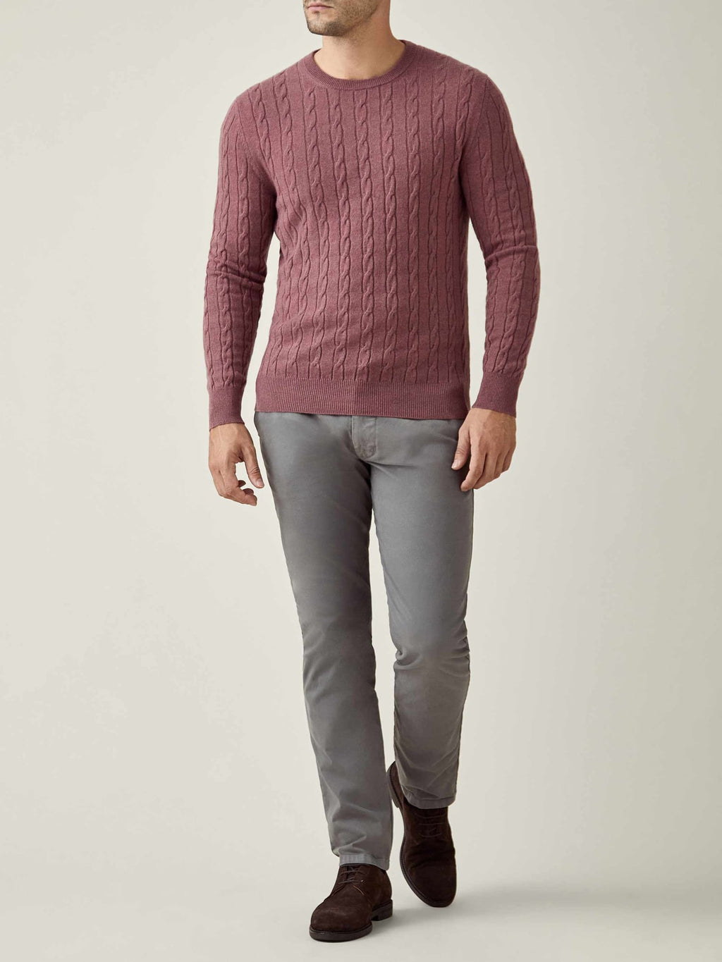 A man models the Luca Faloni Cashmere Cable Knit in Desert Rose, paired with grey trousers for a sophisticated, casual look.