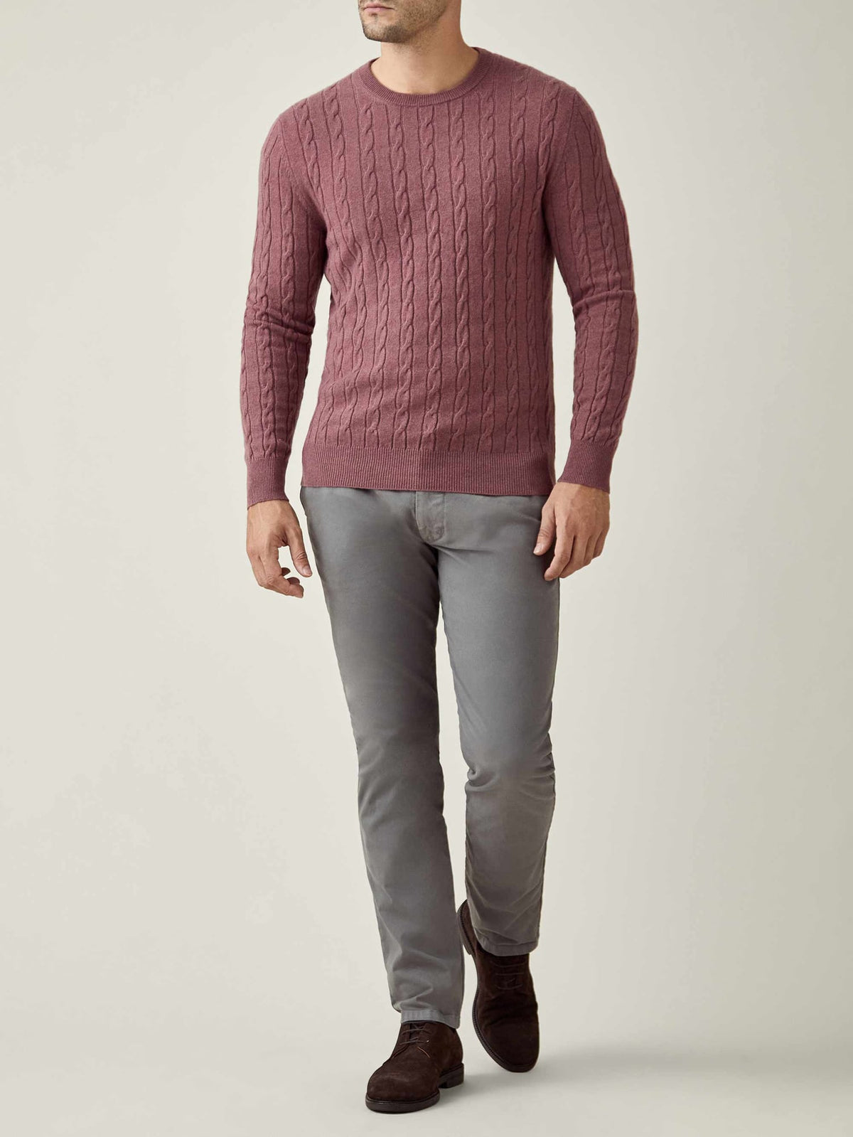 A man models the Luca Faloni Cashmere Cable Knit in Desert Rose, paired with grey trousers for a sophisticated, casual look.