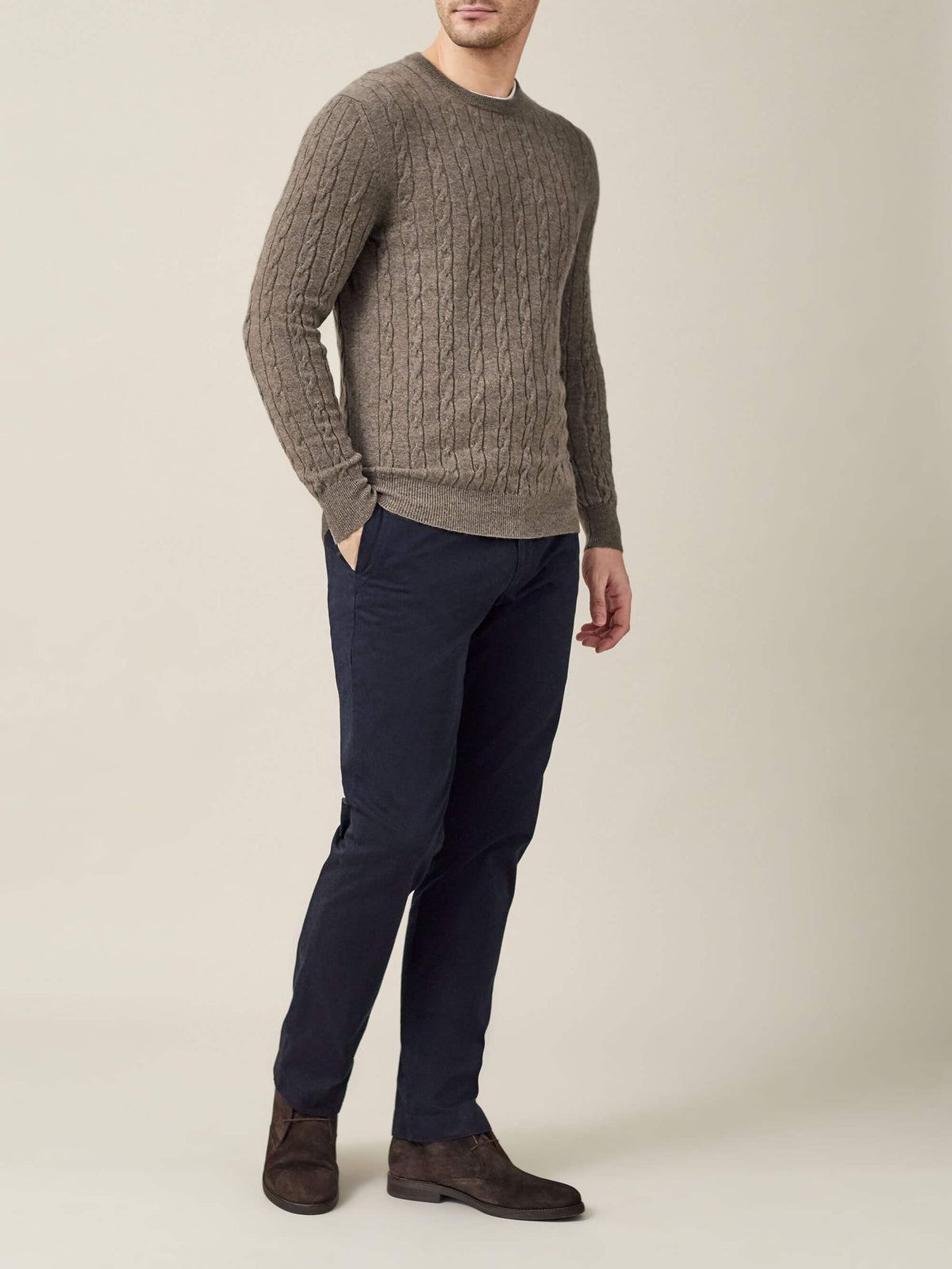 Full length photo of a man wearing a nocciola brown cashmere cable knit jumper, navy chinos and dark brown boots.
