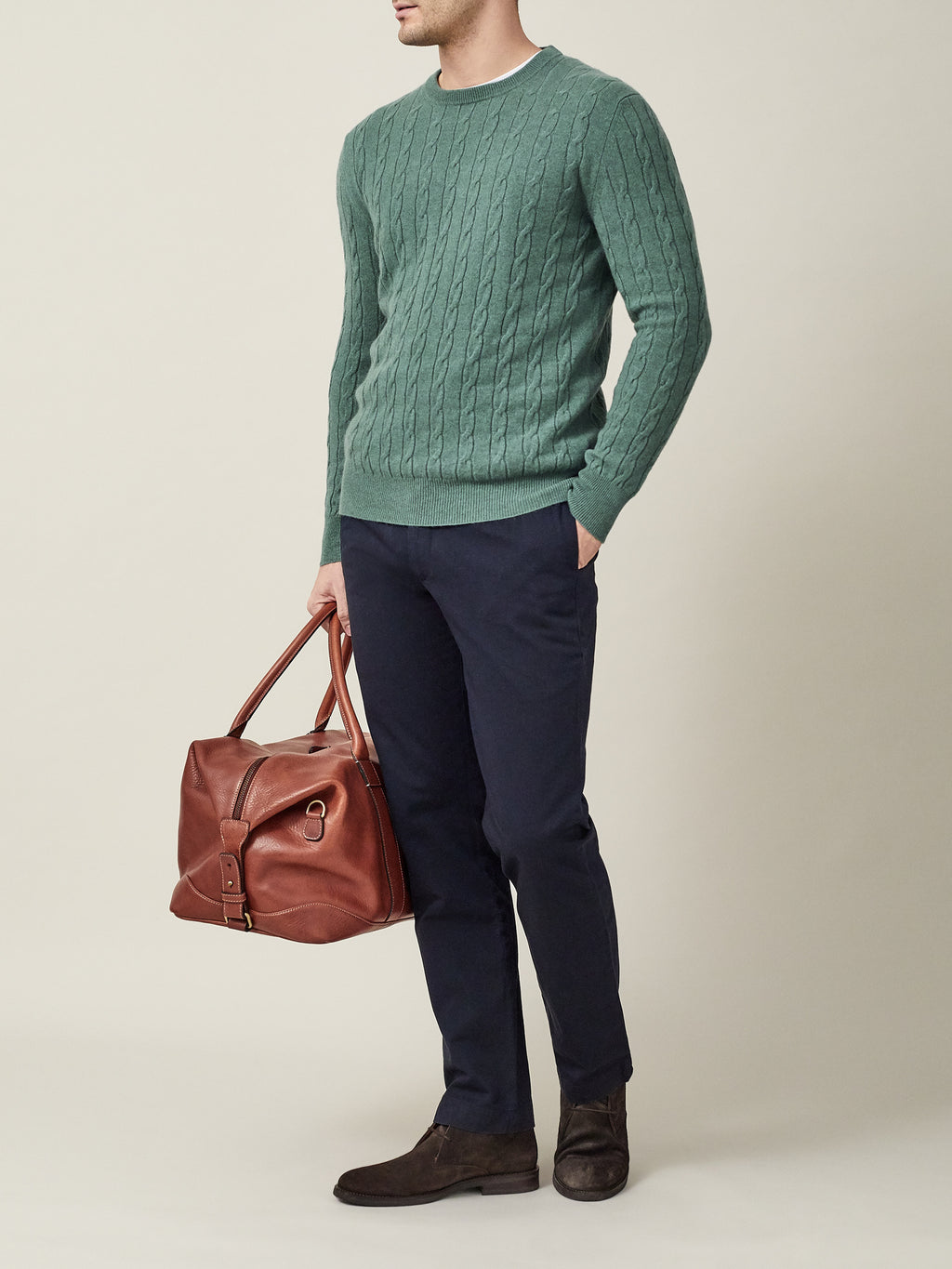 Model styling the marine green cashmere cable knit jumper with navy chinos and holding a brown leather travel bag.