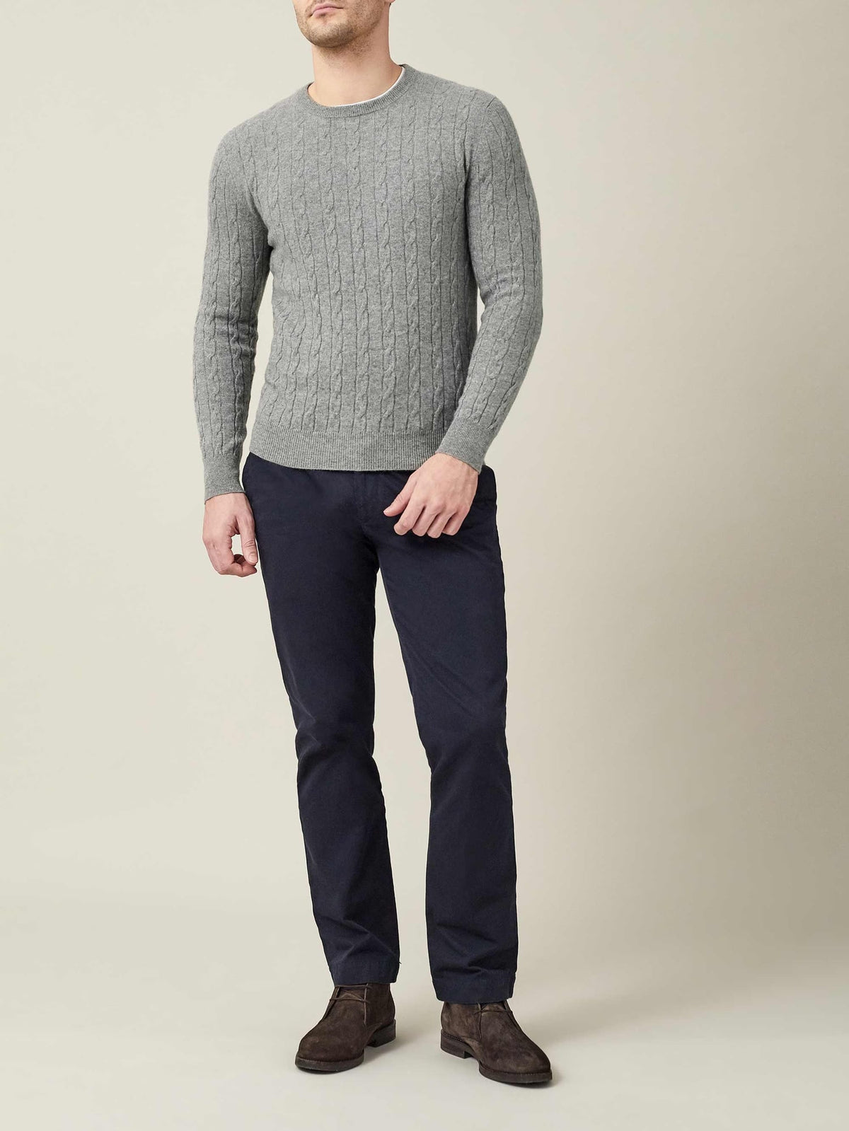 A model stands wearing the Dolomiti Grey cashmere cable knit jumper, paired with dark navy trousers and brown suede boots.