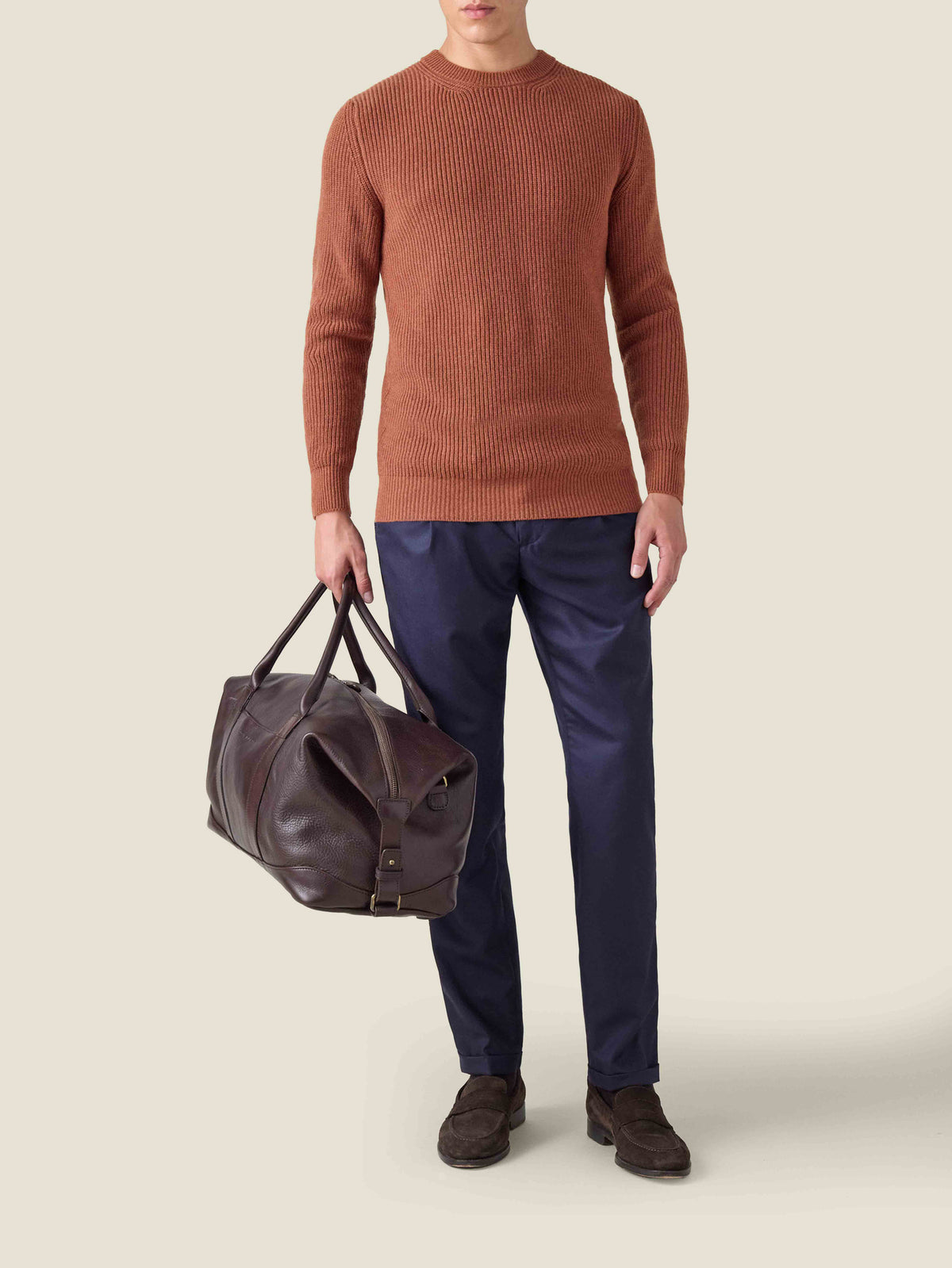 Front view of a model wearing the Chunky Knit Crew Neck in Rust with navy trousers, holding a brown leather travel bag.