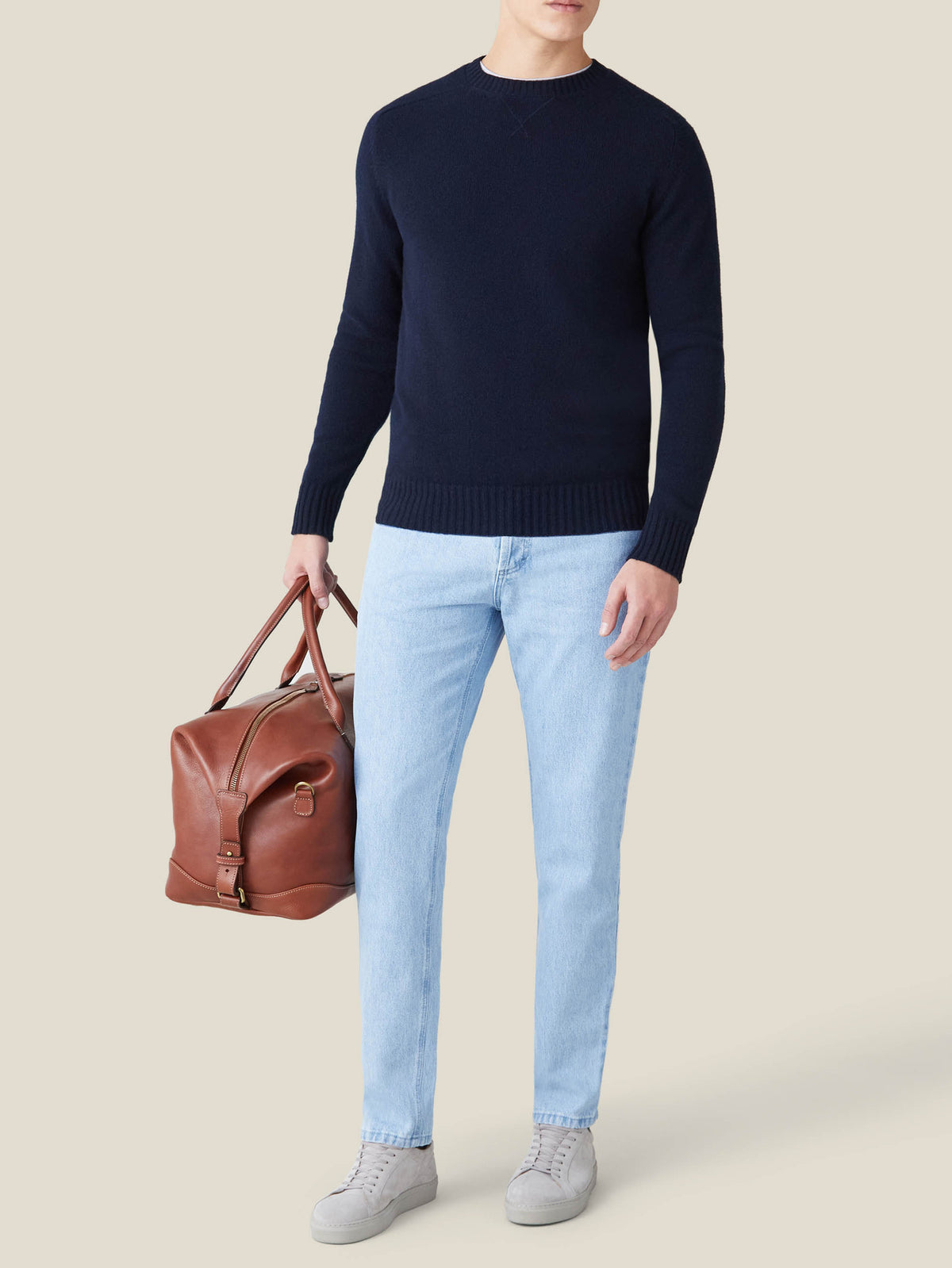 A man wears the Country Crew Neck in midnight blue with light jeans and grey sneakers, holding a brown leather bag.