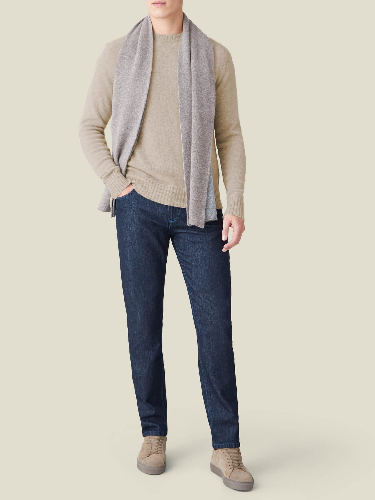 A man models the Camel Beige Country Crew Neck with a grey scarf, dark denim jeans and beige sneakers.