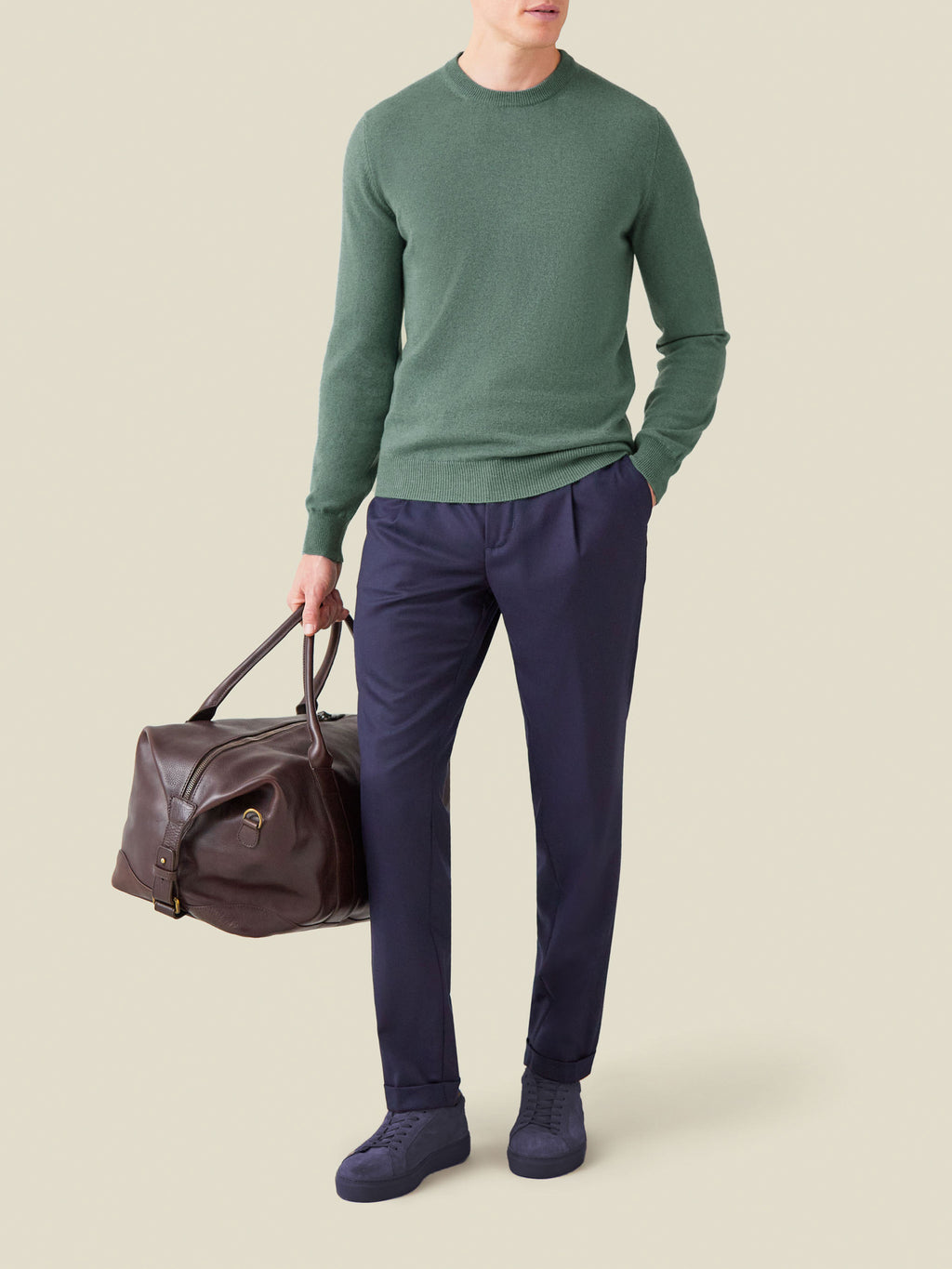 Model wears the Marine Green Cashmere Crew Neck with navy trousers, holding a brown leather duffel bag.