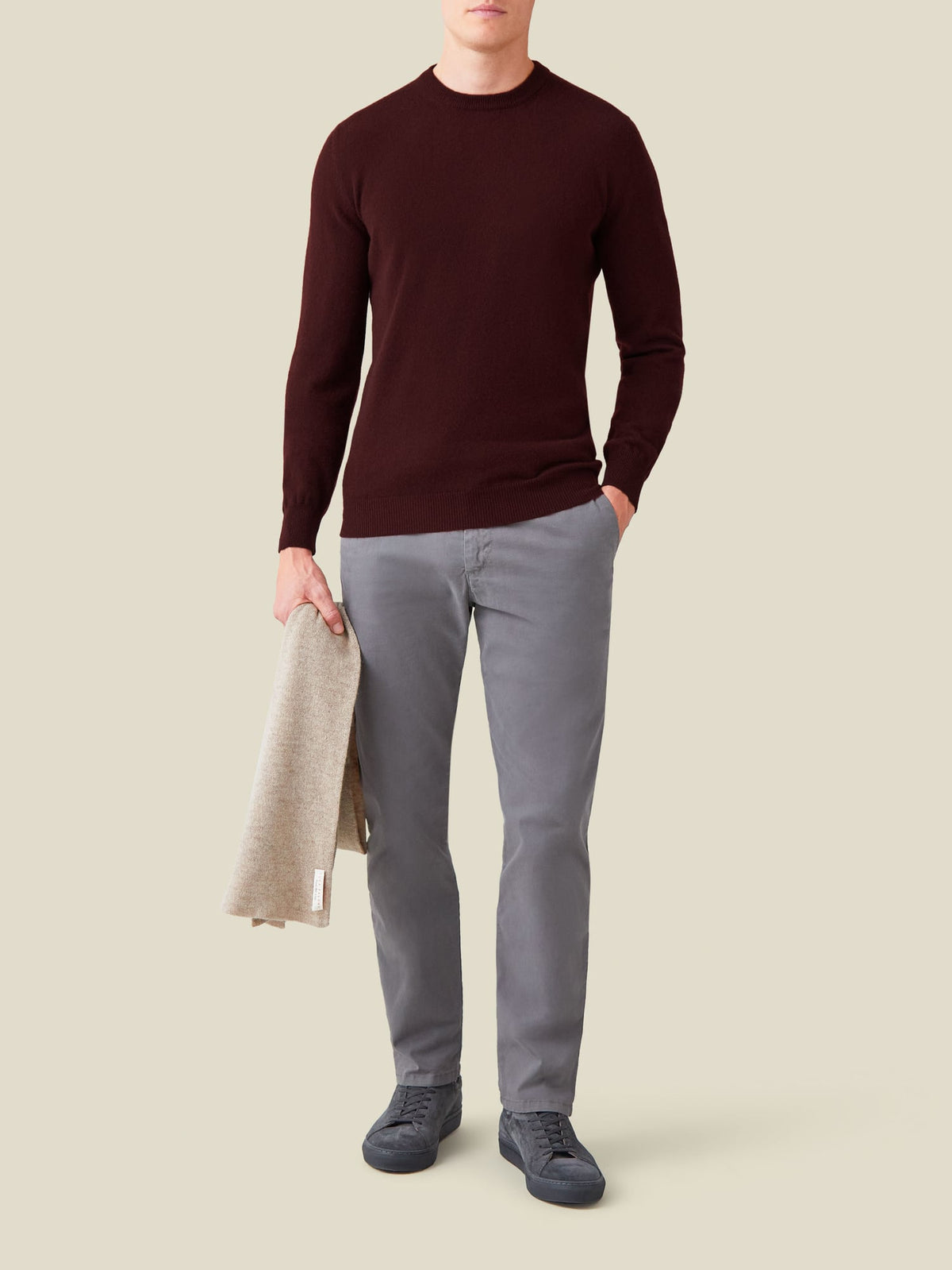 A man wears a lava red cashmere crew neck sweater with grey trousers, holding a beige scarf against a plain background.