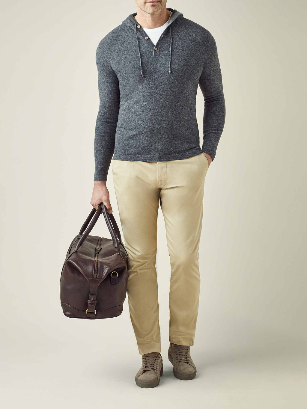 A model wears the Stone Grey Cashmere Hoodie, paired with light brown chinos and holding a dark brown leather duffel bag.