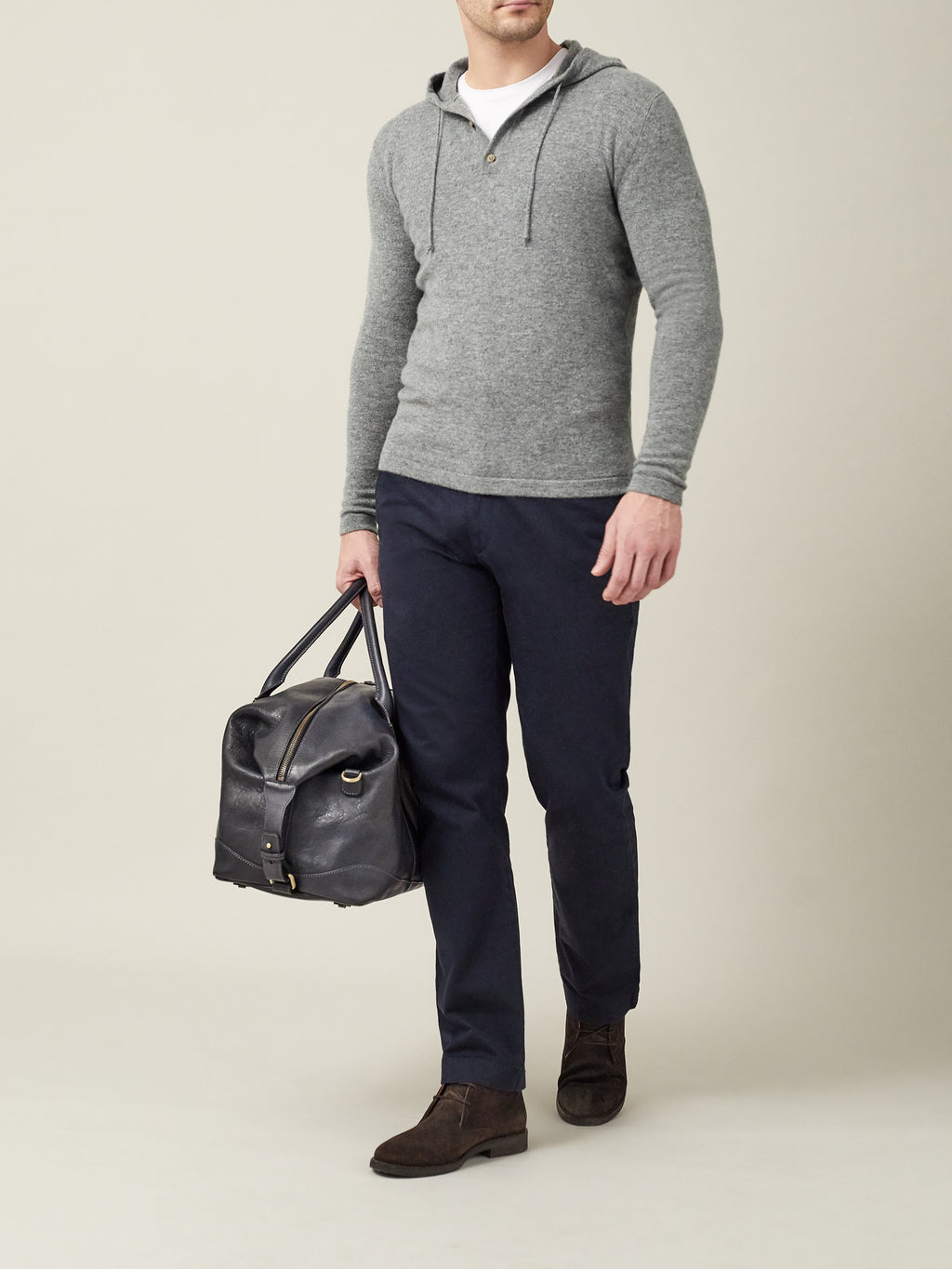A model wears the Luca Faloni Cashmere Hoodie in Dolomiti Grey with navy trousers, brown boots and a leather weekend bag.