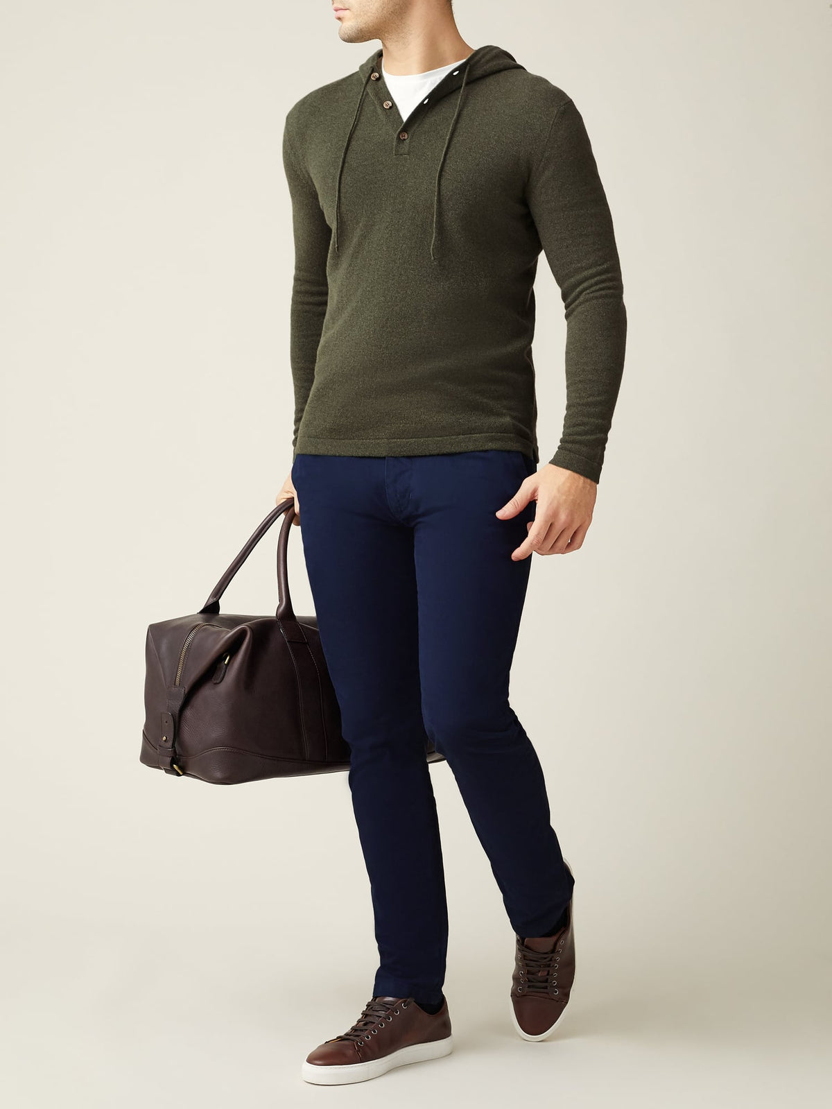 Model in a hunting green cashmere hoodie, navy trousers, and brown trainers, holding a brown leather bag.