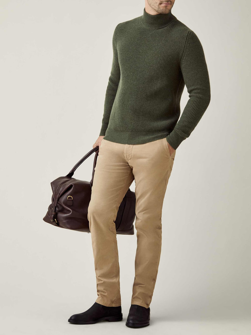 Man models the Chunky Knit Mock Neck in Hunting Green, styled with beige chinos and carrying a brown leather travel bag.