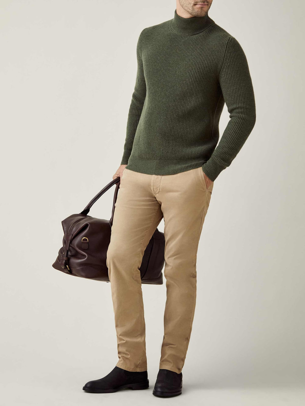 Man models the Chunky Knit Mock Neck in Hunting Green, styled with beige chinos and carrying a brown leather travel bag.