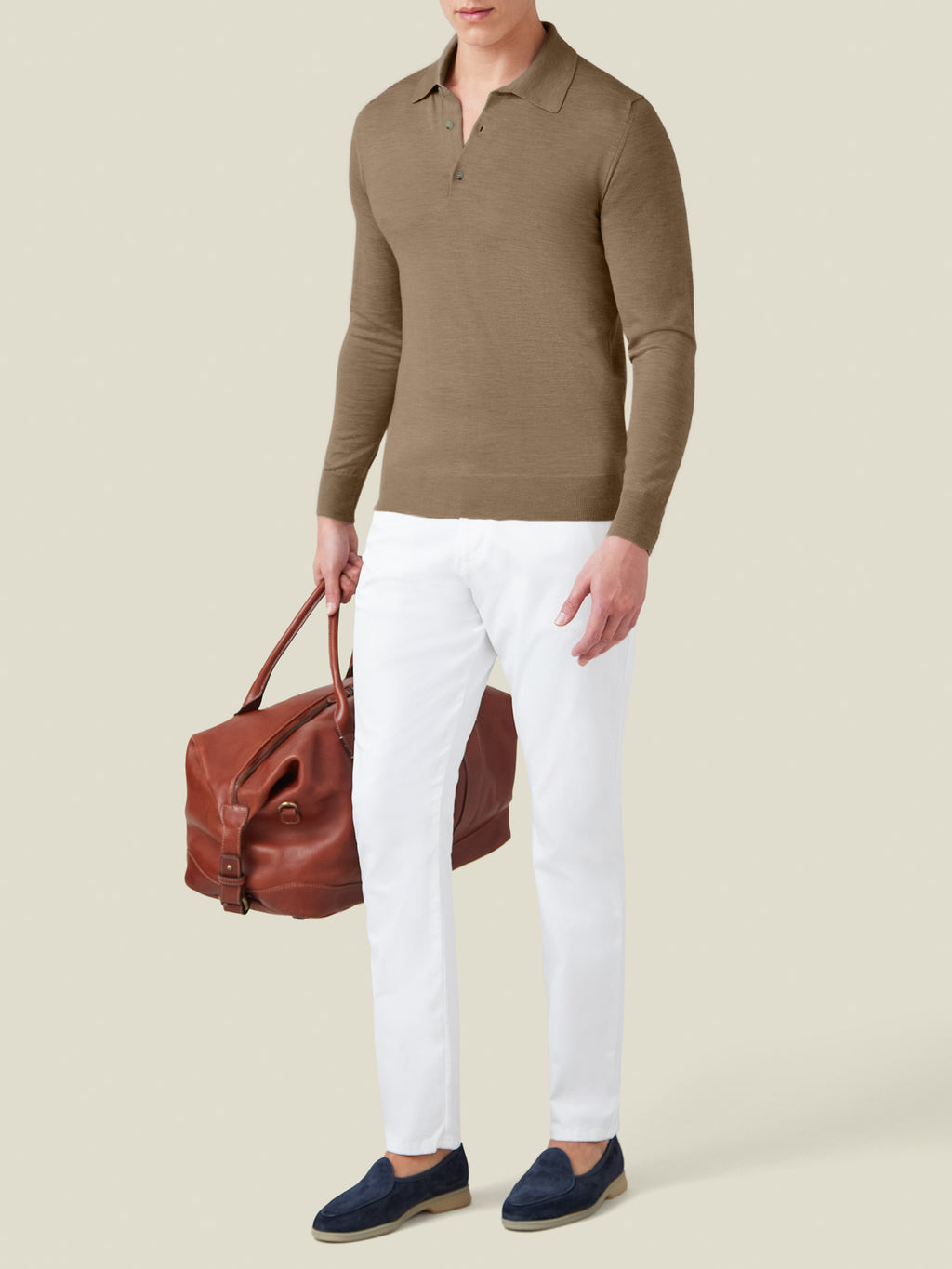 Model wears the Fine Silk Cashmere Polo in Caramel Brown, paired with white trousers and a brown leather holdall bag.