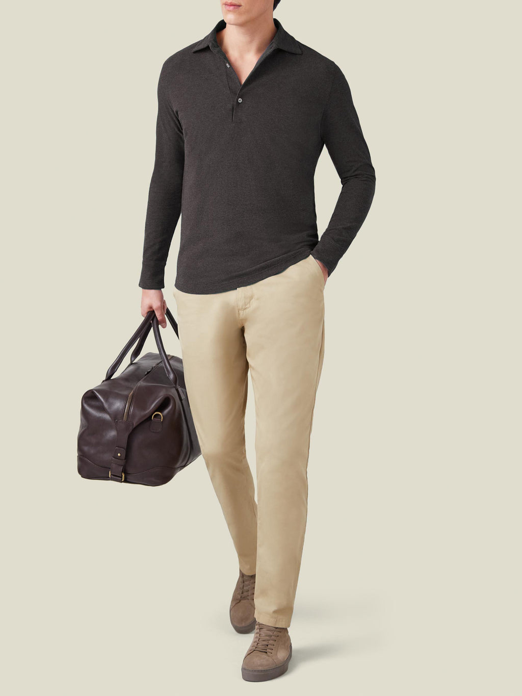 Model wears the Charcoal Grey Brera Piqué Polo Shirt with beige chinos and holds a brown leather travel bag.