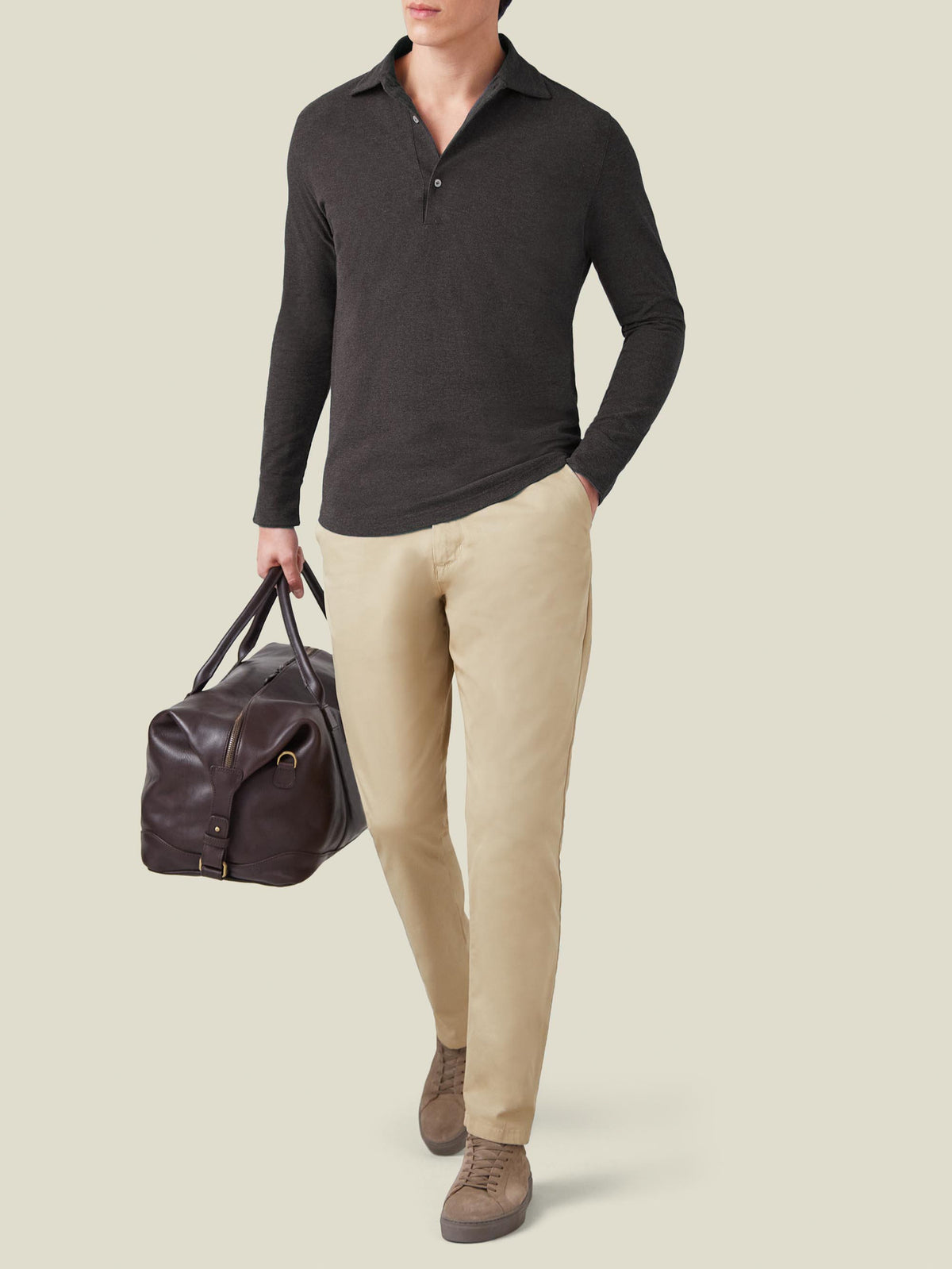 Model wears the Charcoal Grey Brera Piqué Polo Shirt with beige chinos and holds a brown leather travel bag.
