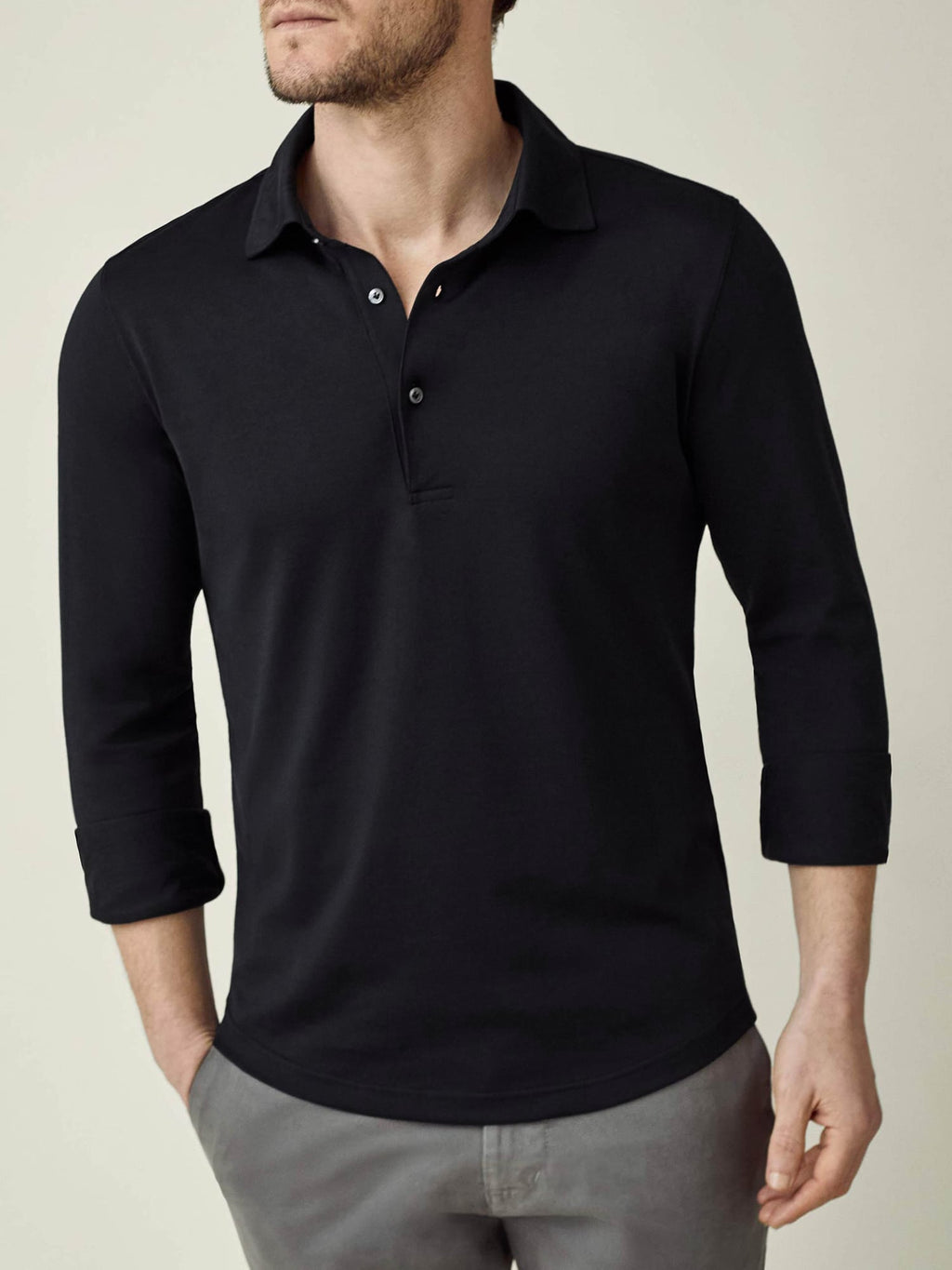 A man models the black long-sleeved Amalfi Silk-Cotton Polo Shirt, styled with rolled-up sleeves and grey trousers.
