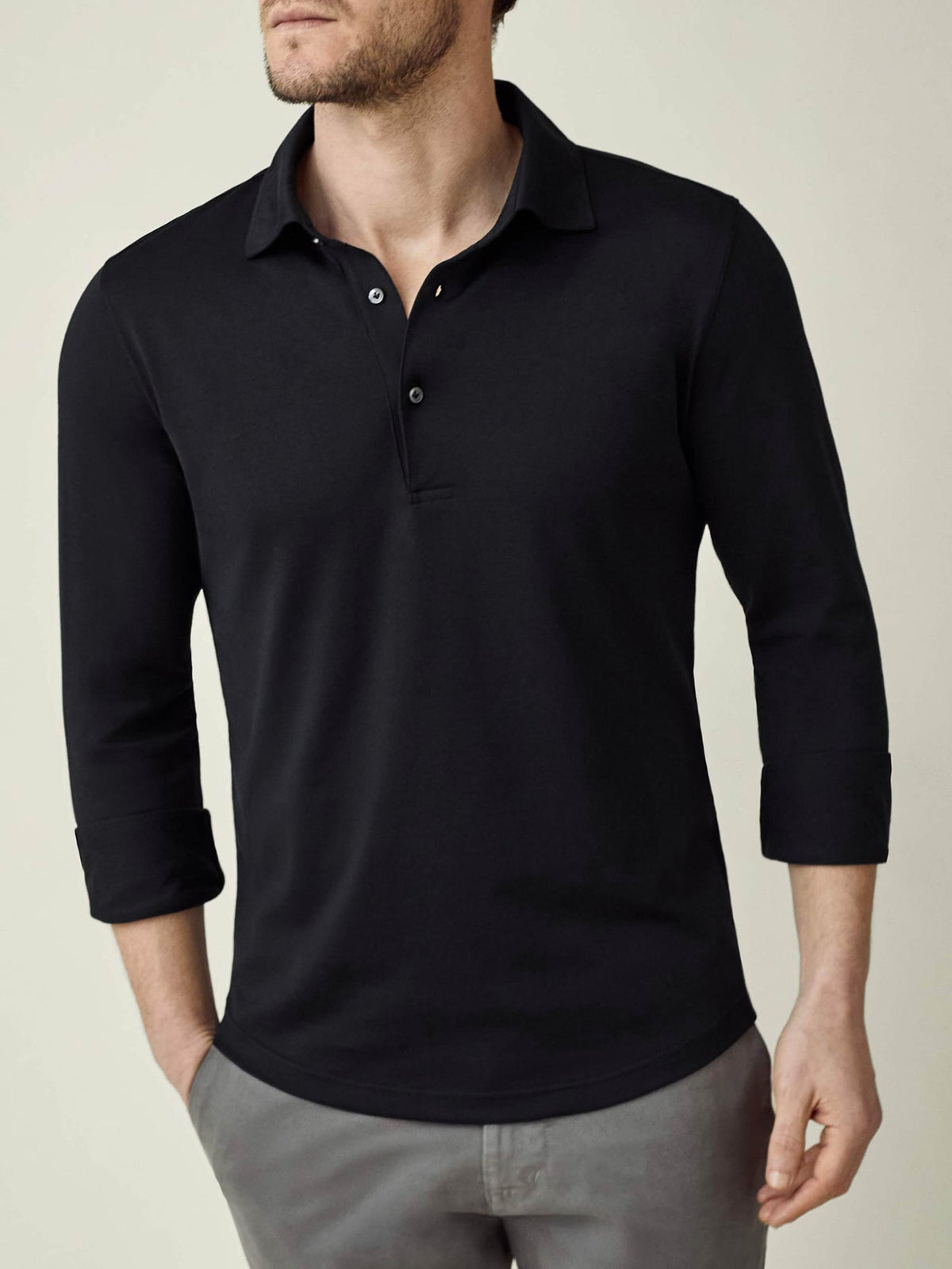 A man models the black long-sleeved Amalfi Silk-Cotton Polo Shirt, styled with rolled-up sleeves and grey trousers.