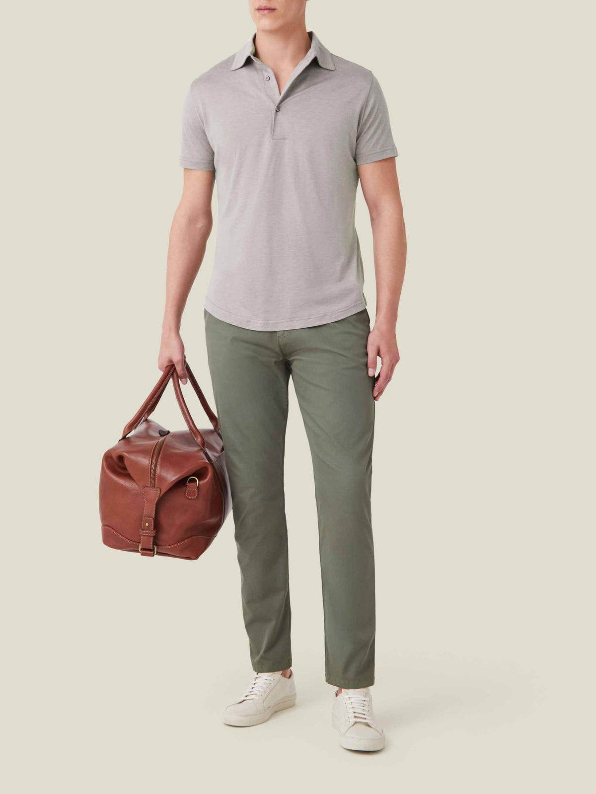 Model wears the Nocciola Brown Silk-Cotton Polo with olive chinos and white sneakers, holding a brown leather weekender bag.