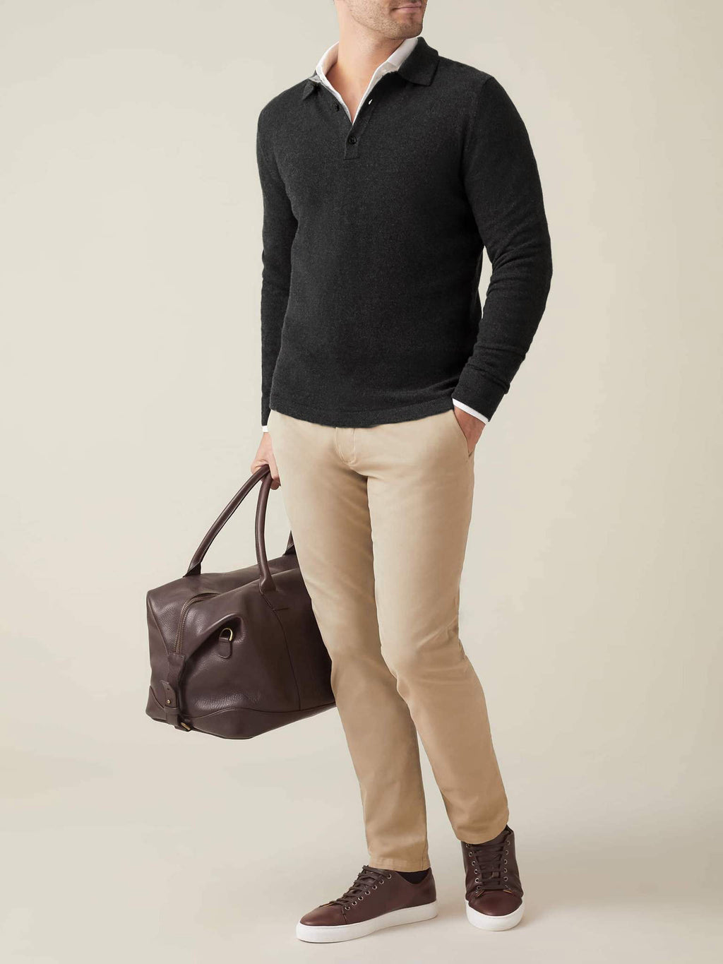 Man wearing a charcoal grey cashmere polo sweater over a white shirt, with beige chinos and holding a brown travel bag.