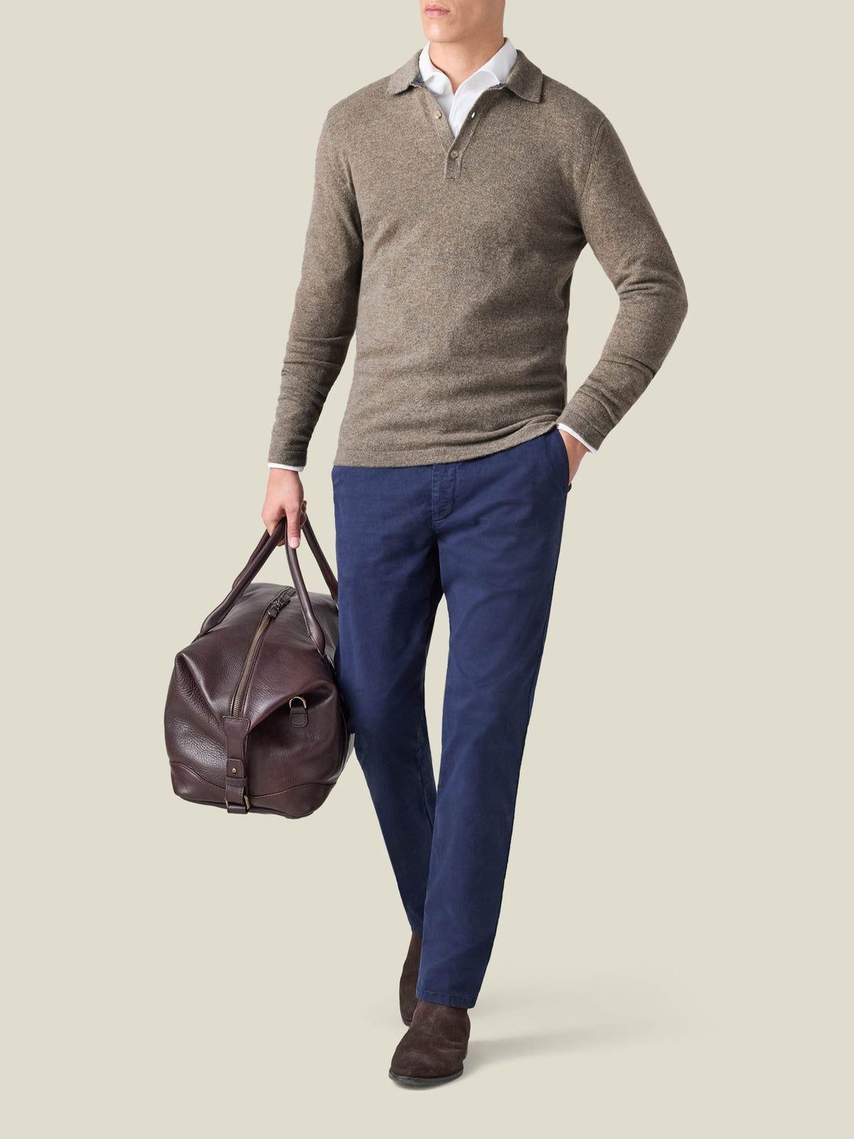 A man wears the Nocciola Brown Cashmere Polo Sweater with blue trousers and carries a brown leather travel bag.