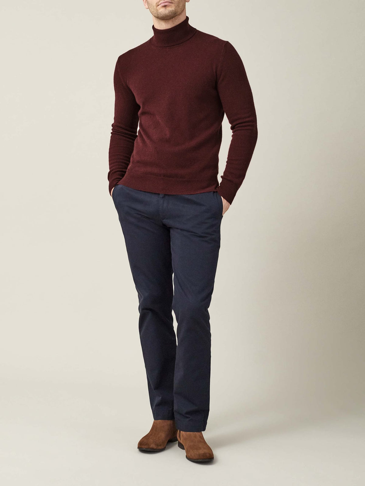 A man models the Luca Faloni Lava Red Cashmere Roll Neck, styled with navy trousers and brown suede boots.