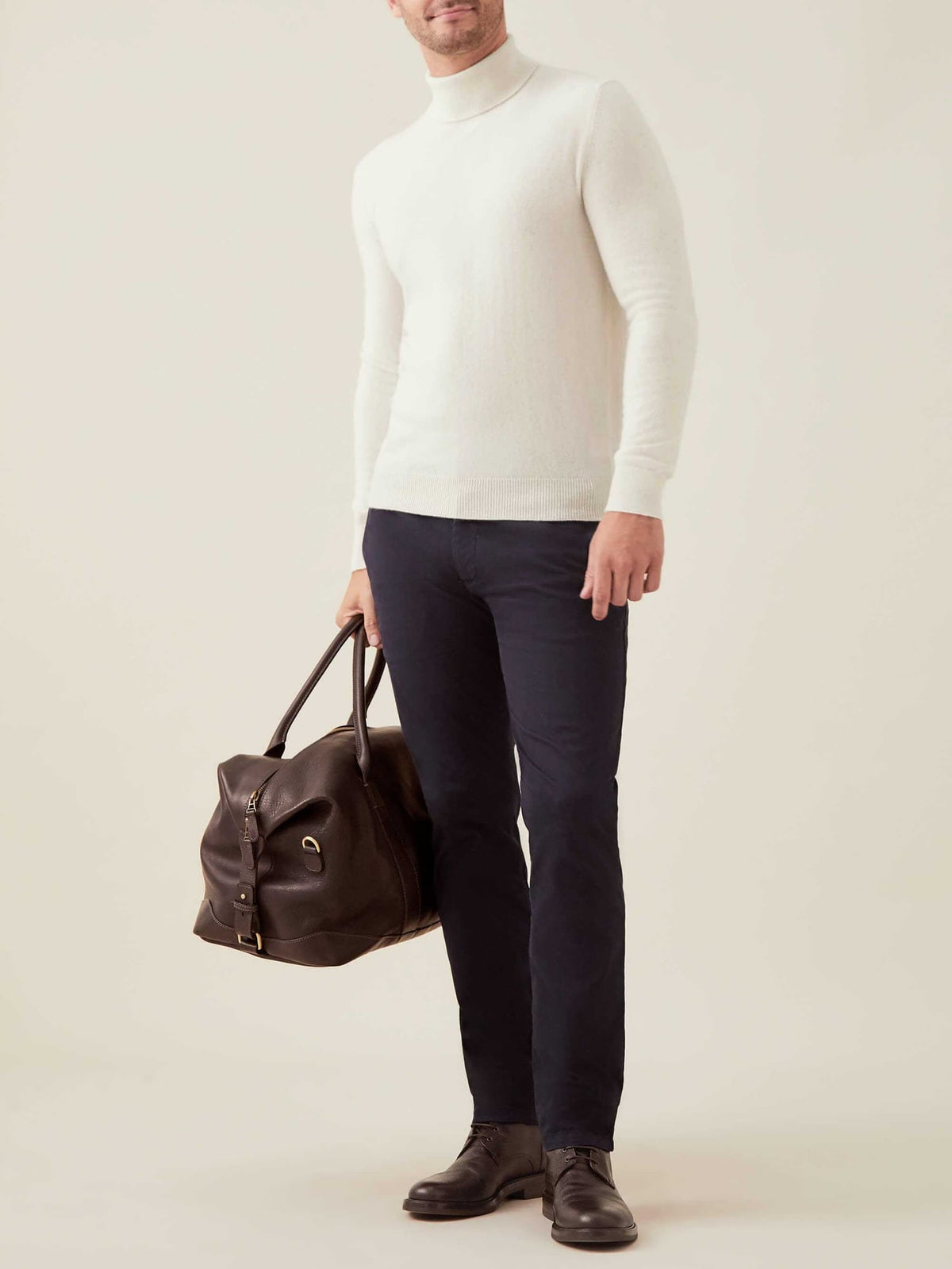 A man models the ivory cashmere roll neck sweater with dark trousers and brown boots, holding a leather travel bag.