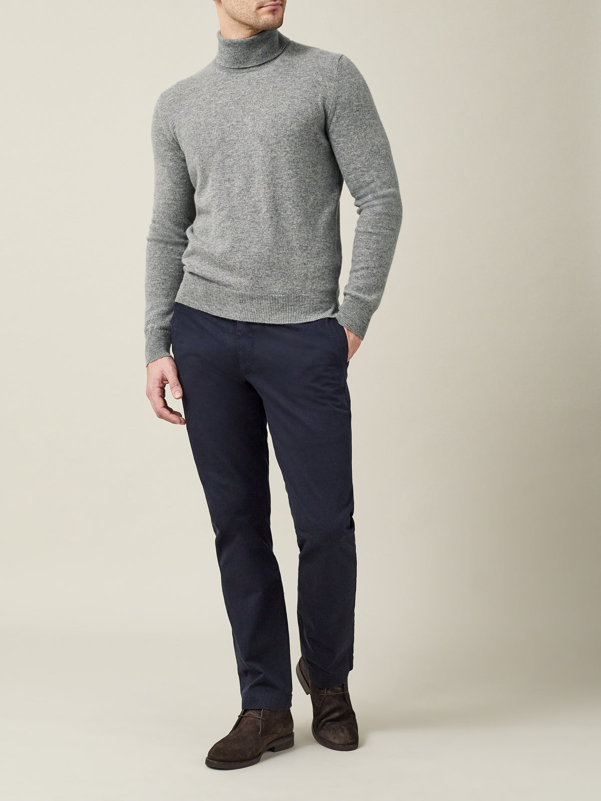 Man wears a Dolomiti Grey cashmere roll neck with dark navy trousers and brown suede boots, pictured from the front.