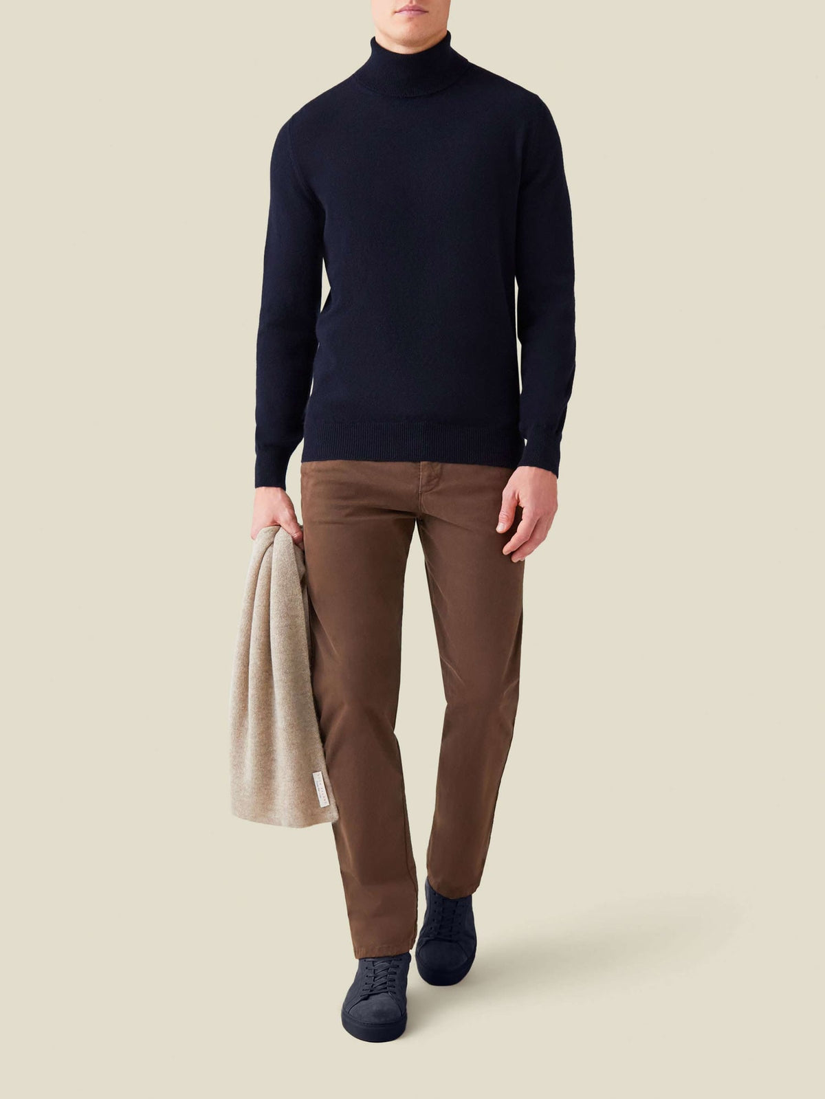 A man presents a full look wearing the midnight blue cashmere roll neck with brown trousers and carrying a beige scarf.