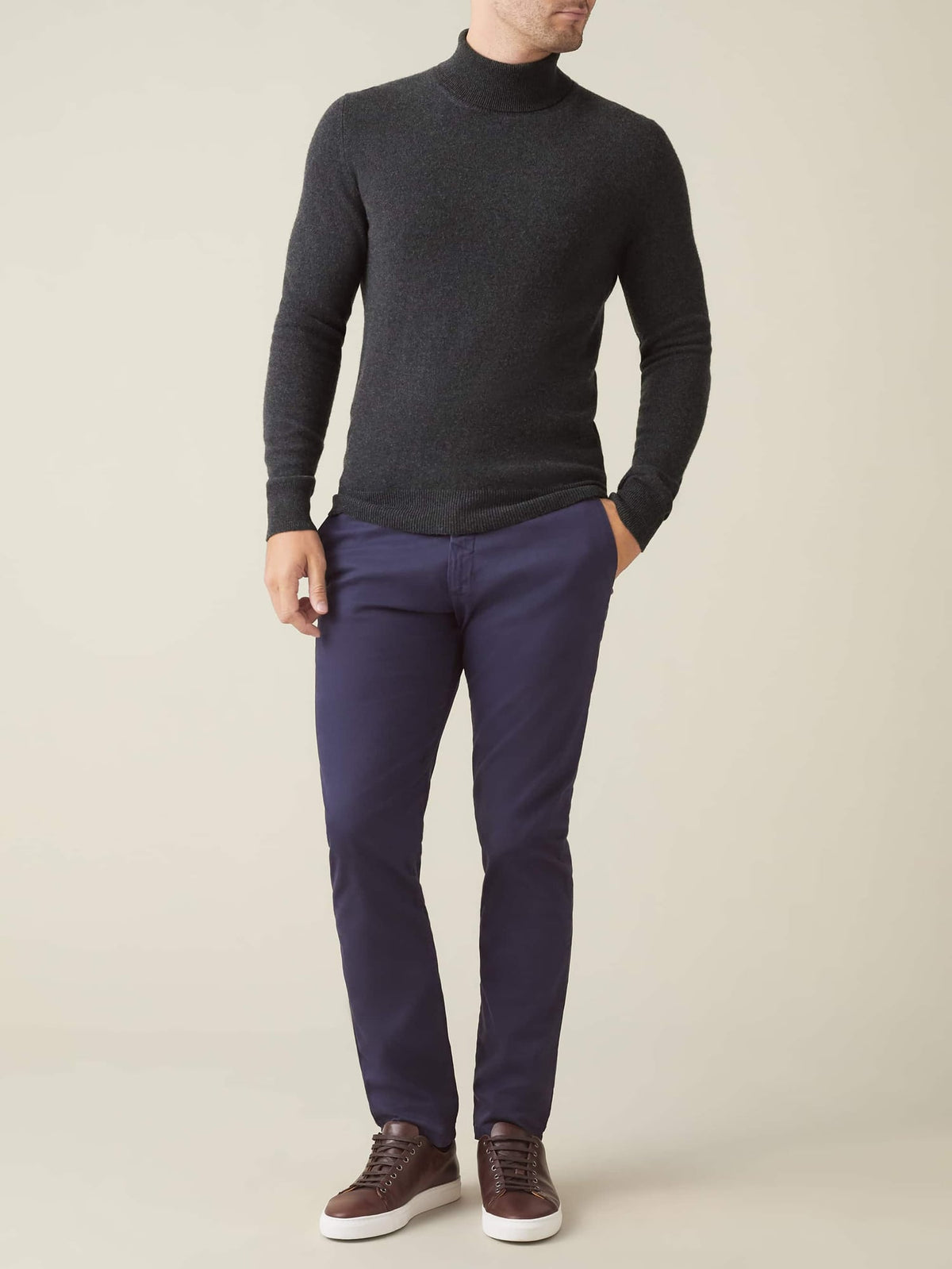 Full-length photo of a man wearing a charcoal grey cashmere roll neck sweater with navy blue trousers and brown sneakers.