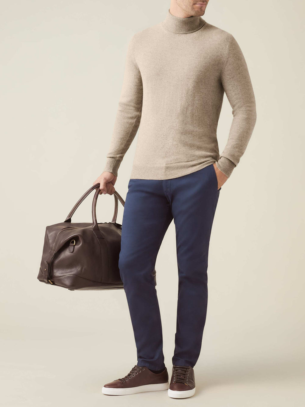 A man models a camel beige cashmere roll neck with navy trousers and brown leather sneakers, holding a brown travel bag.