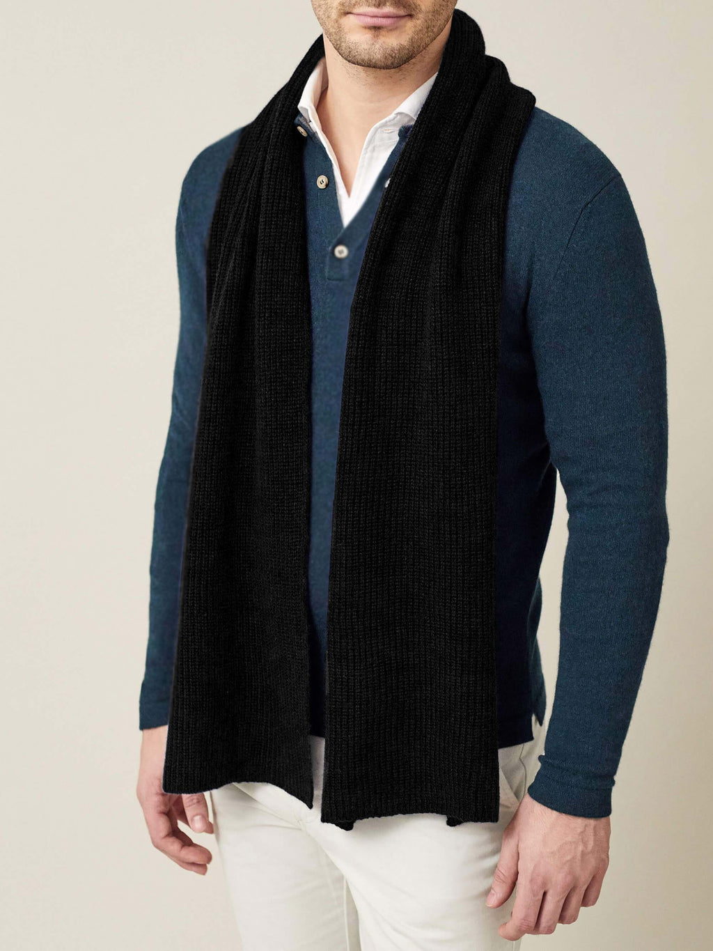 A front view of a model wearing the black chunky knit cashmere scarf over a blue long-sleeve polo and white trousers.
