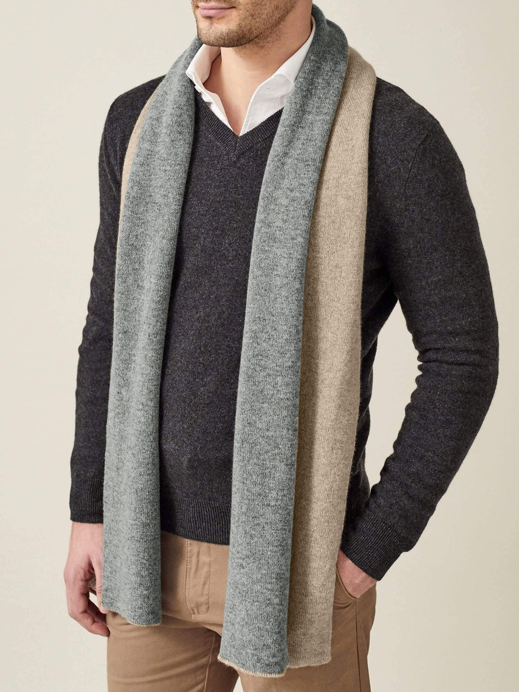 A man models the Double-Faced Cashmere Scarf in camel and grey, styled over a dark grey v-neck sweater.