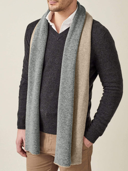 A man models the Double-Faced Cashmere Scarf in camel and grey, styled over a dark grey v-neck sweater.