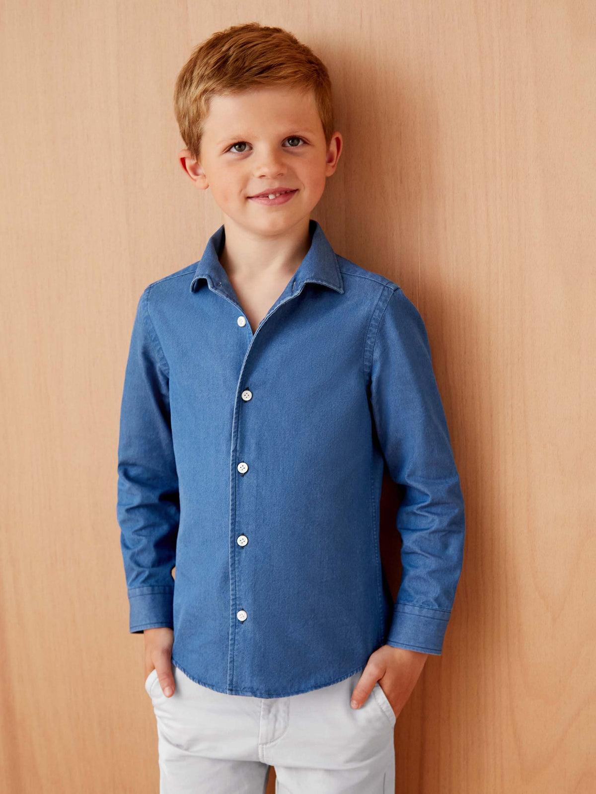 A young boy models the Denim Junior Shirt in Classic Blue with his hands in the pockets of his white trousers.