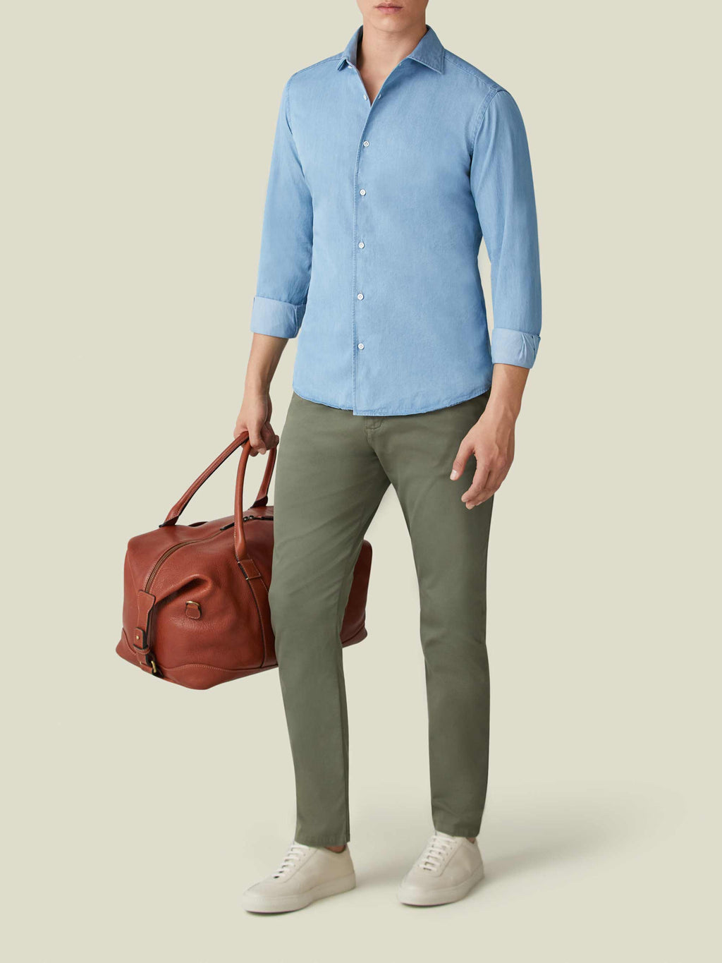 A model wears the Luca Faloni light blue denim shirt with green chinos and white trainers, holding a brown leather bag.