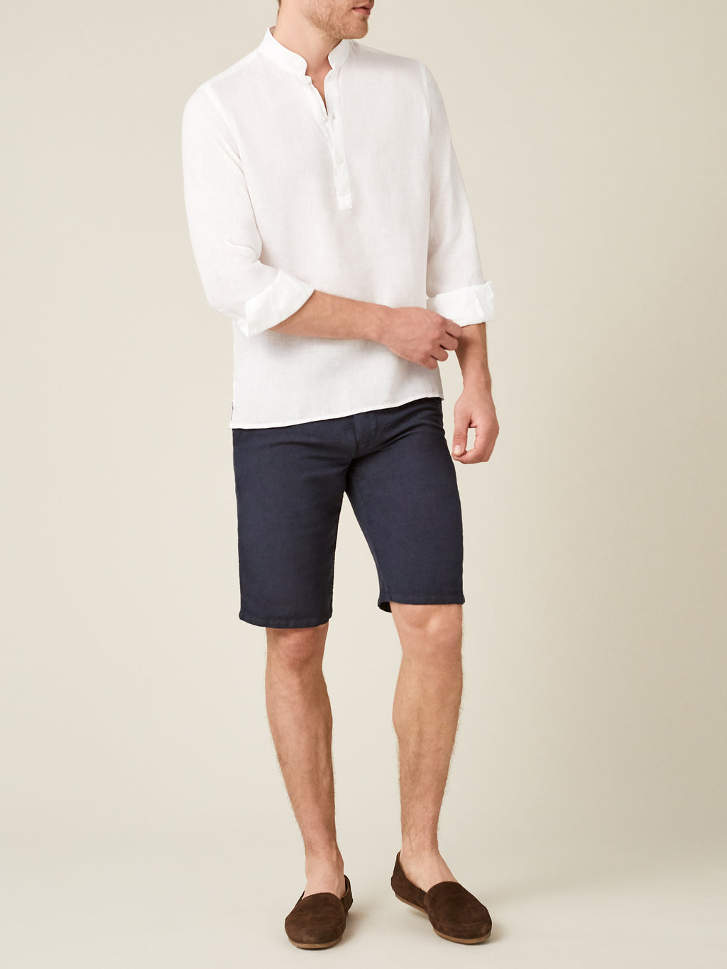 A man wears the white Forte Linen Shirt with dark shorts and brown loafers, rolling up one sleeve.