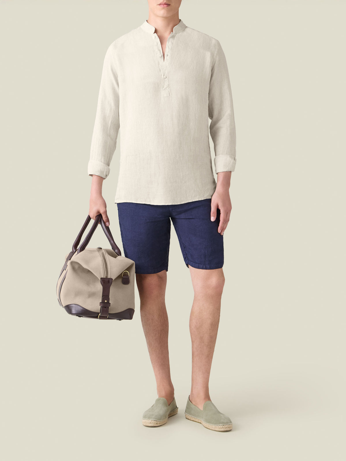 A man wearing the sand Forte Linen Shirt styled with navy shorts and espadrilles, holding a weekend bag.