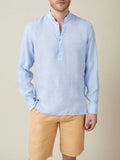 A man models the Forte Linen Shirt in Sky Blue, a popover style with a band collar, styled with light brown shorts.