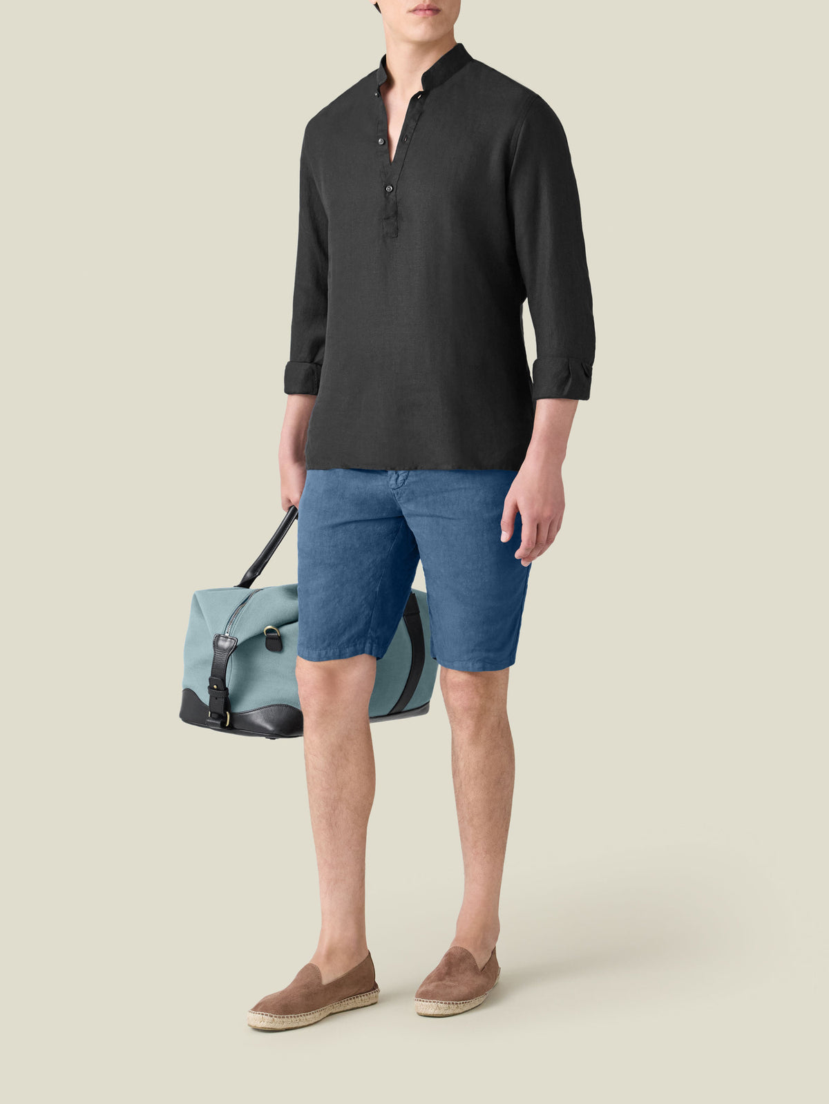 Model wears the black Forte Linen Shirt with blue shorts and brown espadrilles, holding a light blue duffle bag.