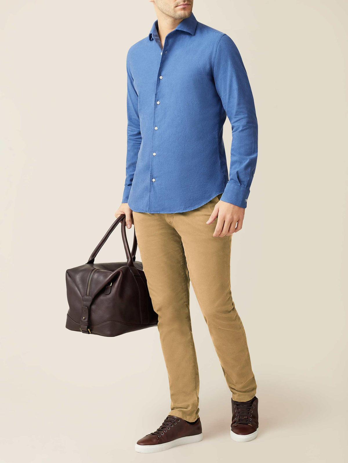 Model wears the Brushed Cotton Shirt in Chambray Blue, styled with tan chinos and holding a brown leather travel bag.