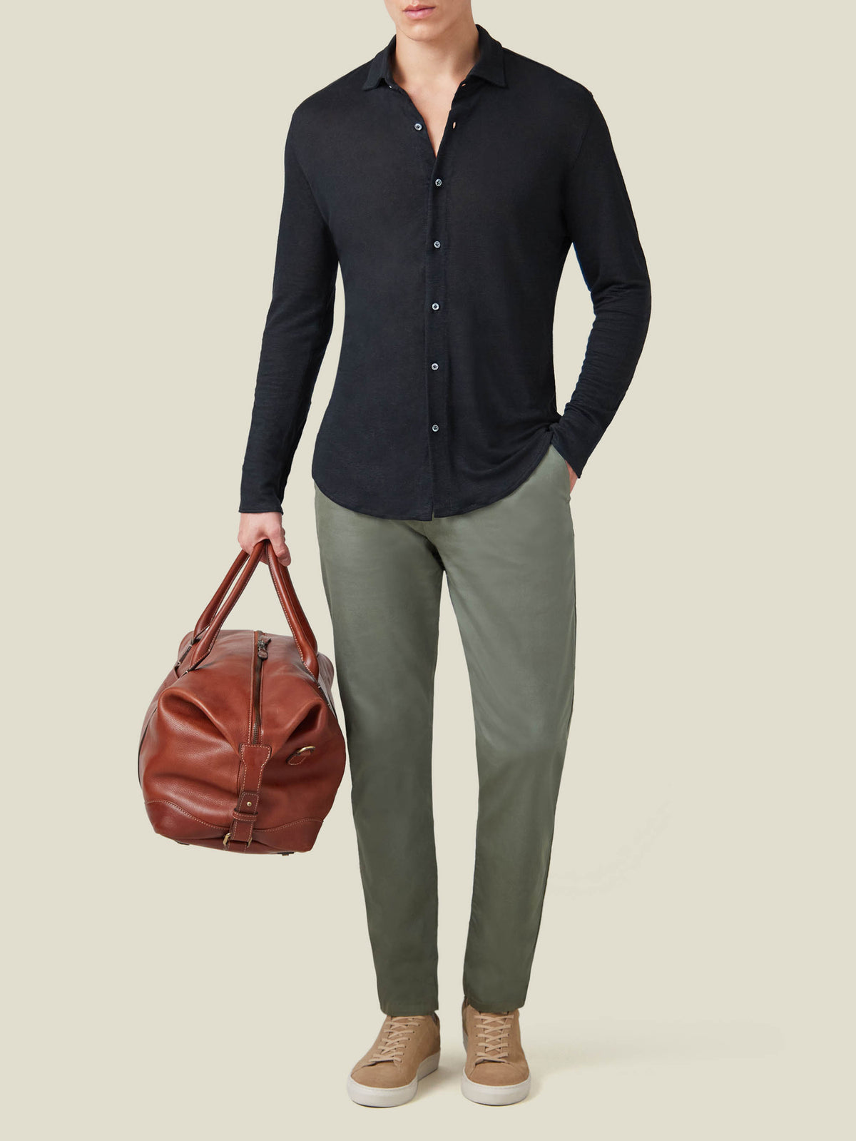 Model wearing a black linen jersey shirt, olive green trousers, and light brown sneakers, holding a brown leather bag.