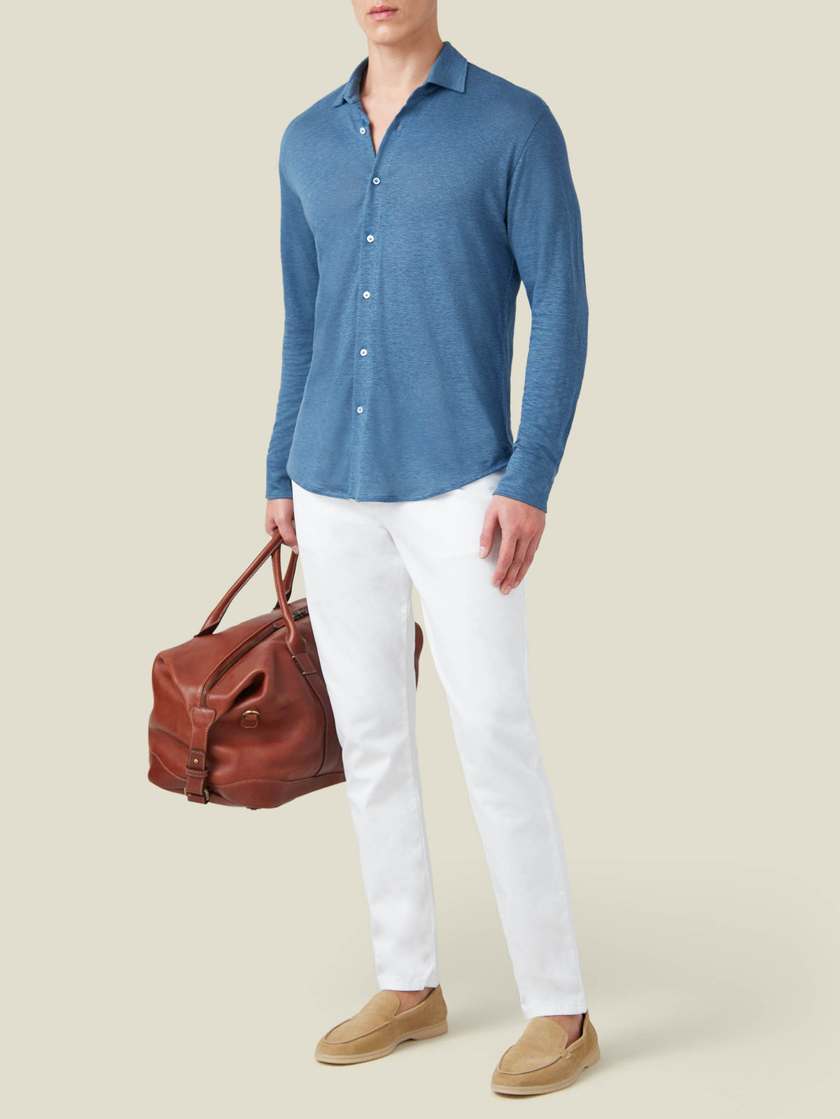 Model wears the Steel Blue Linen Jersey Shirt with white trousers and loafers, holding a brown leather travel bag.