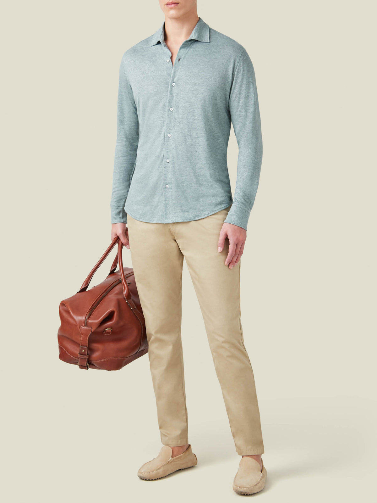 Man wearing the Sea Grey Linen Jersey Shirt with light trousers and loafers, holding a brown leather travel bag.