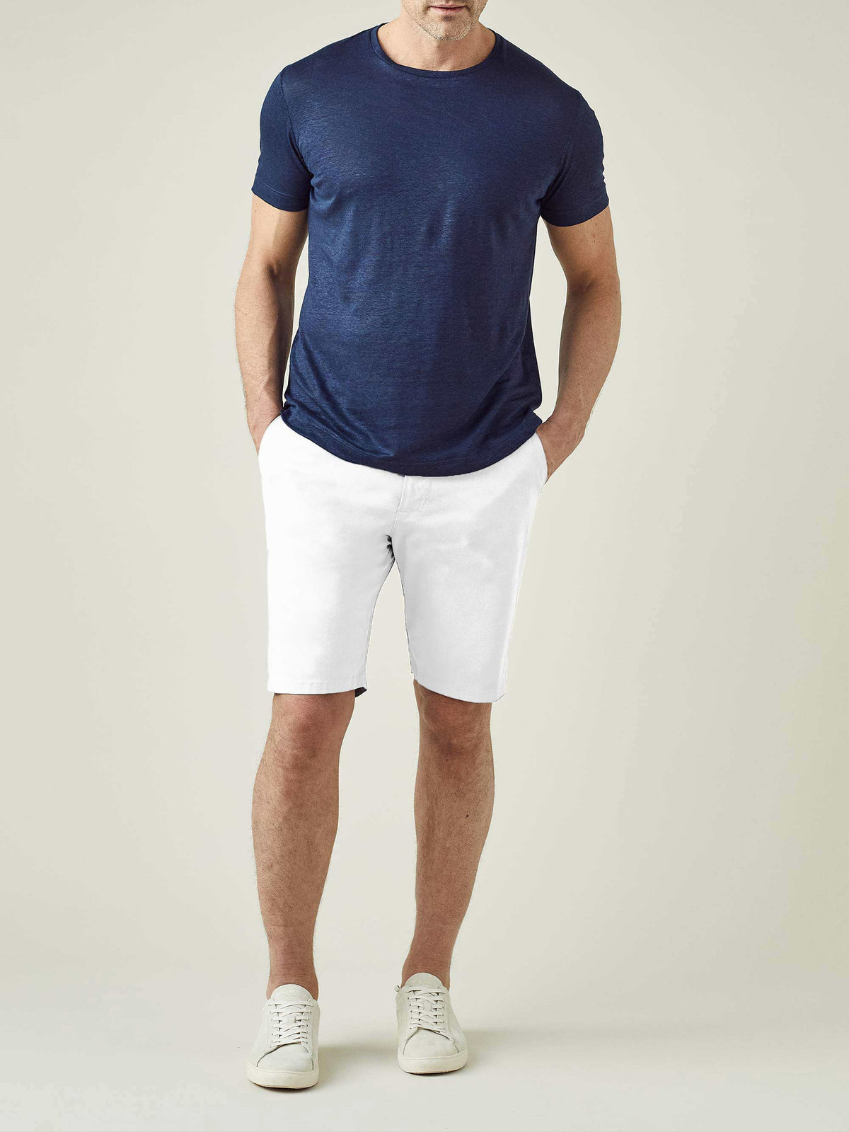 A man models the Luca Faloni White Cotton Shorts, styled with a navy t-shirt and white sneakers, in a full-length front view.