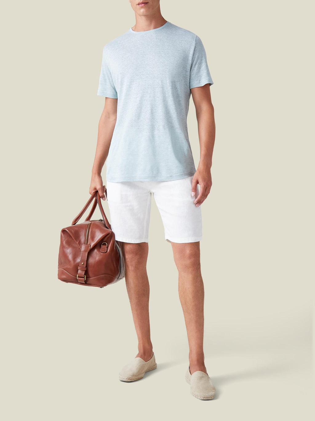 Model wears white Bermuda Linen Shorts, a light blue t-shirt, cream espadrilles, and holds a brown leather bag.